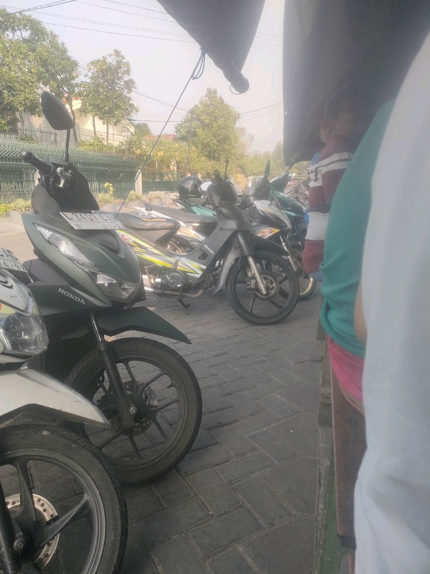 The image depicts a street scene in Surabaya, Indonesia, with several parked motorcycles. The foreground features the front of two motorcycles, one dark green and the other silver, with their wheels and handlebars visible. The dark green motorcycle has "HONDA" branding on its side panel and a license plate that reads "L 2101 CA." Behind these, a line of other motorcycles is parked, including a green one with yellow and black decals and a black one with a helmet on its seat. The motorcycles are parked on a brick-paved sidewalk. In the background, there are trees and buildings, and a portion of a black tarp or awning hangs over the scene from the top right. A person wearing a striped shirt, red shorts, and a turquoise shirt is partially visible on the right side of the image, standing near the motorcycles. The overall lighting suggests it is daytime, possibly late afternoon, given the hazy sky.