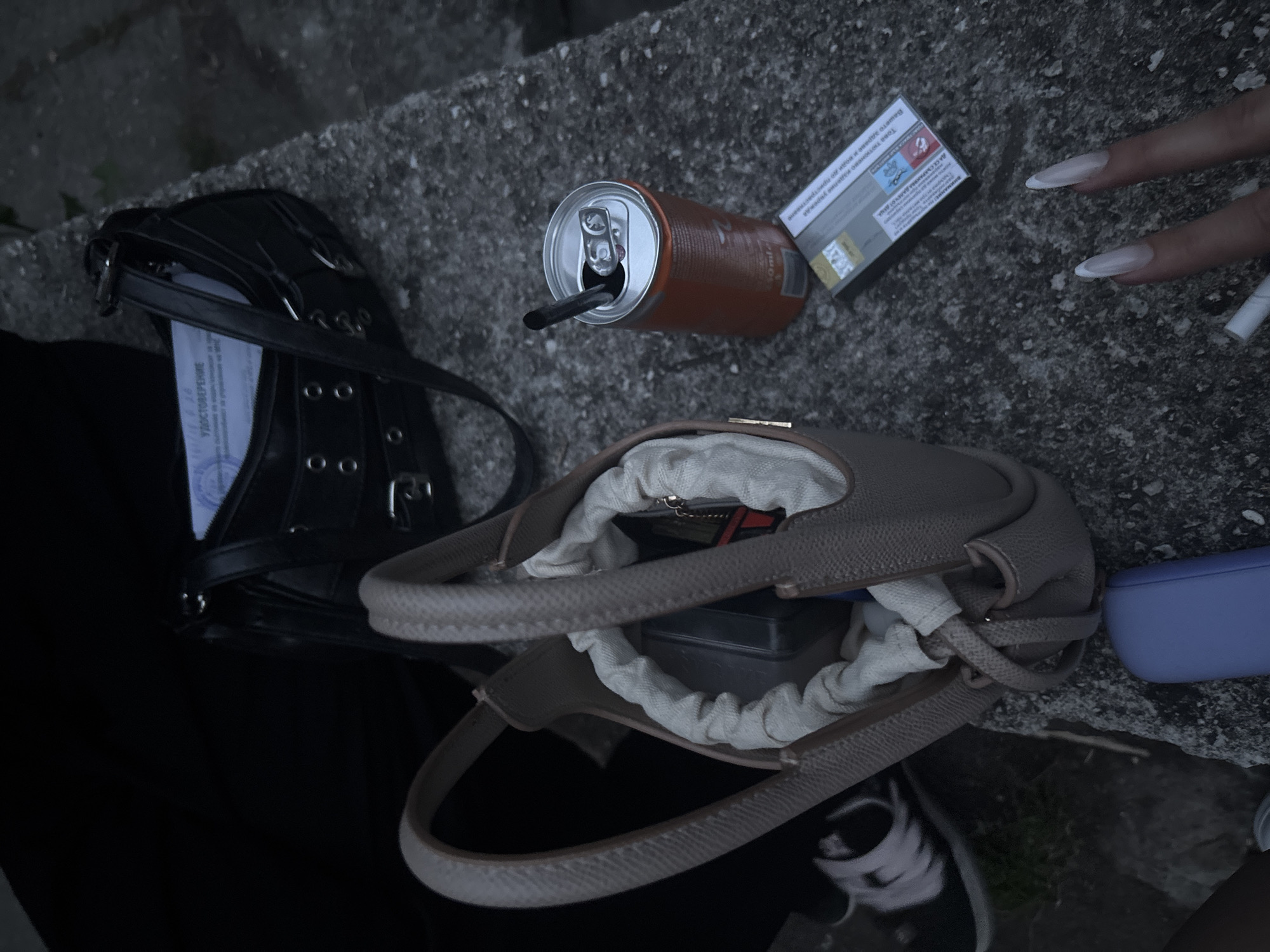 The image depicts an outdoor scene at dusk, possibly on a concrete ledge in Plovdiv, Bulgaria, given the Cyrillic text visible on a document and the cigarette pack. In the foreground, a woman's hand with long, white-tipped fingernails is visible, reaching towards a cigarette. A cigarette pack, appearing to be of a European brand with Cyrillic warning labels, lies on the concrete next to an open, orange soda can with a straw in it.
Behind these items are two handbags. A black, slightly worn shoulder bag with silver buckle details sits open, revealing a document with Cyrillic text. Next to it, a smaller, light-colored crossbody bag is placed, its contents partially visible, including a dark rectangular object and a keyring with a charm. To the left of the bags, a person's leg wearing a black shoe with white laces is visible, suggesting the presence of at least one person. The overall lighting is dim, indicating evening or overcast conditions. The background shows a dark, out-of-focus area, possibly foliage.
