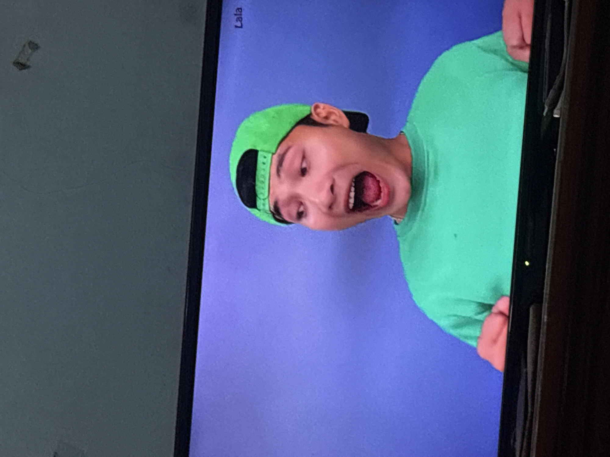 The image shows a young man with a surprised or excited expression, mouth open, wearing a bright green t-shirt and a green baseball cap with a black brim. He is positioned in the center of the frame against a solid purple background. His hands are visible at the bottom of the frame, suggesting he is leaning forward or interacting with something out of view. In the top right corner of the screen, the word "Lala" is visible. The image appears to be a screenshot or a photograph of a screen displaying content, as indicated by the texture and faint reflections on the screen surface. The context of Palimanan, Indonesia, is not discernible from the visual information presented in the image itself.