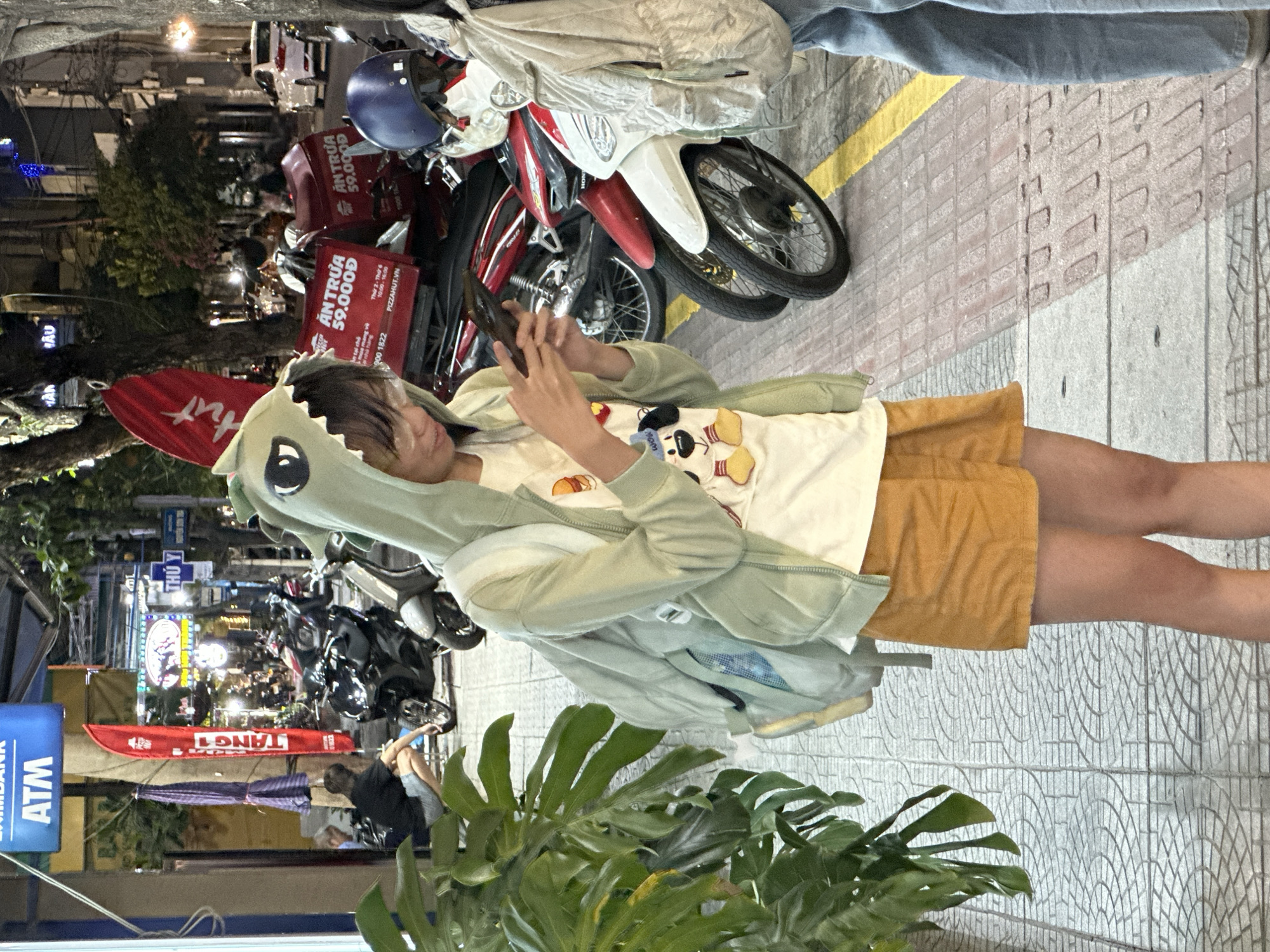 A young person wearing a light green dinosaur-themed hoodie and an orange skirt is standing on a sidewalk in Da Nang, Vietnam, looking at their phone. They have a backpack slung over their shoulder. In the background, several motorcycles are parked along the street, some with large red delivery boxes on the back. A sign for "ATM" is visible on a pole. The sidewalk is paved with patterned tiles. A large green plant is in the foreground. The scene appears to be a commercial or residential street at night, with artificial lighting from streetlights and shop signs.