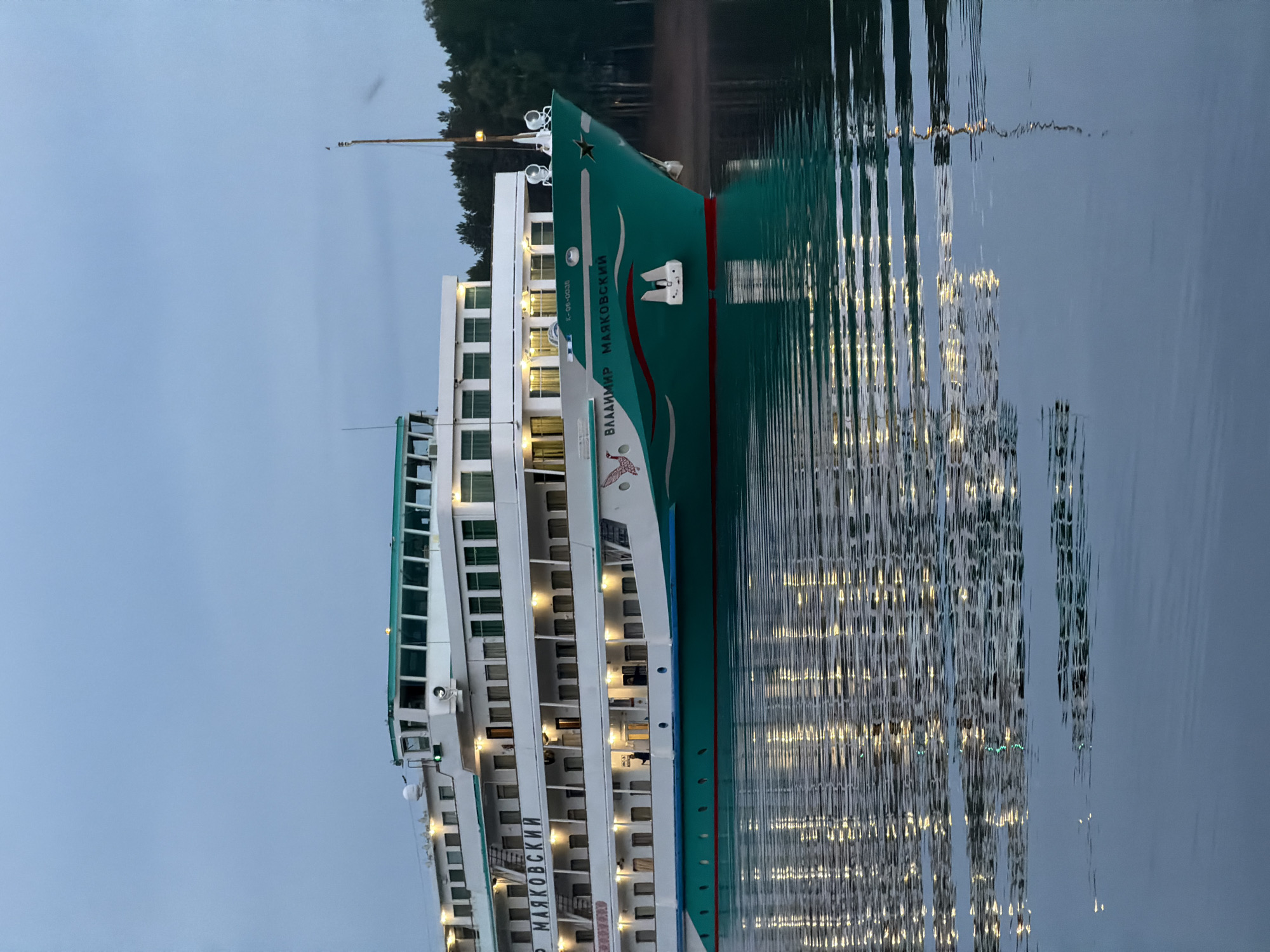 The image shows a large, green and white river cruise ship named "Vladimir Mayakovsky" docked in what appears to be a river. The ship has multiple decks with lit windows. The hull of the ship is green with red and white accents. The ship's reflection is visible in the calm water. Trees line the far shore in the background under an overcast sky. Based on the name of the ship being Cyrillic, it is very likely in Russia, potentially in the city of Chaikovsky, as that is a port located on the Kama River, and a logical place for river cruise ships. There are no people visible in the frame.