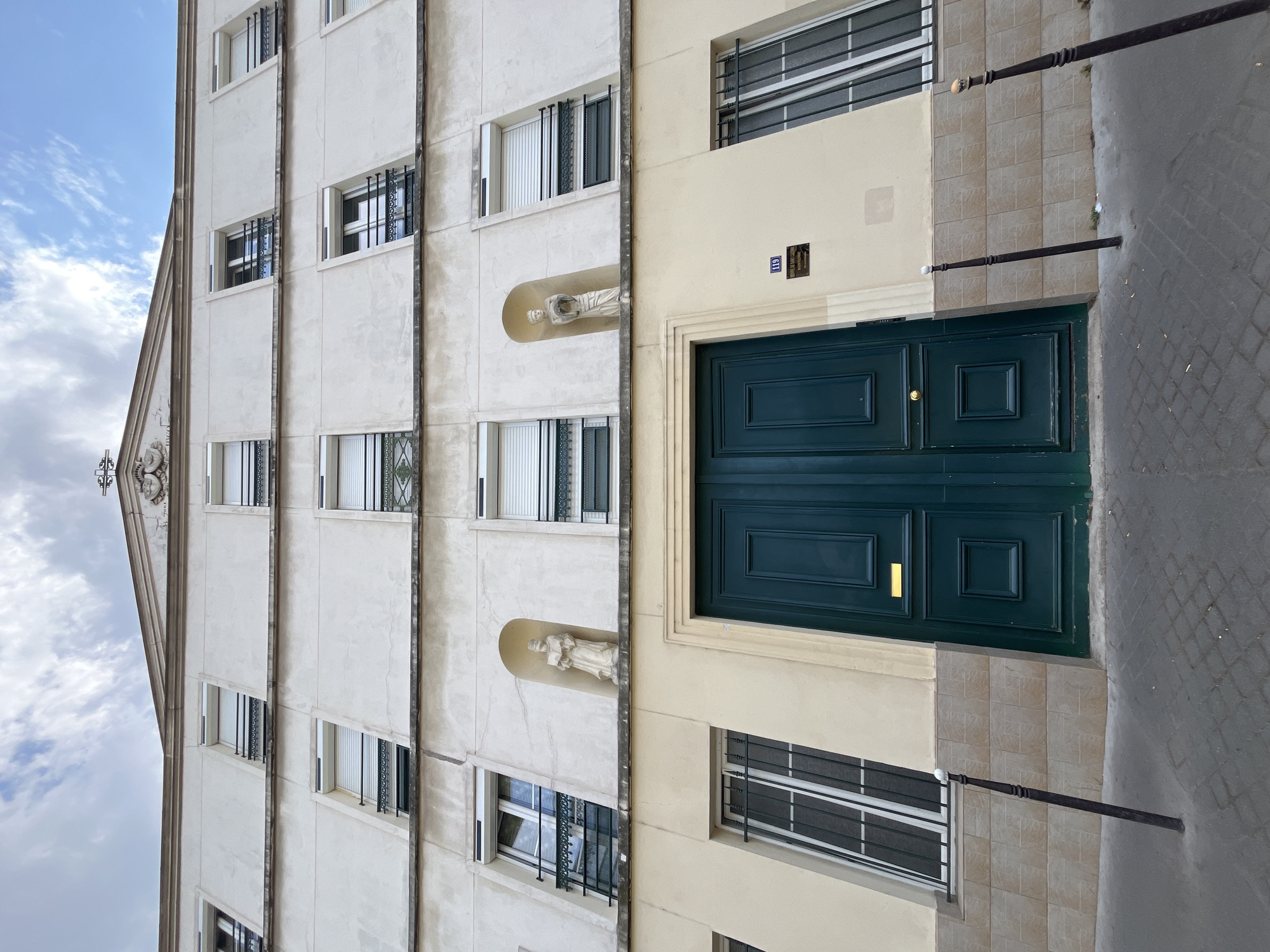 This is an image of a building with a dark green double door. The building has a light-colored facade with several windows, some of which have metal bars. There are two statues of figures in niches on the facade, one above the other. Above the door, there is a sign with the number "119" and some text. The building appears to be located on a street, with a sidewalk made of cobblestones. The sky is visible in the upper left corner of the image and appears to be cloudy. The lighting suggests it is daytime. The overall scene is one of an exterior architectural view of a building.