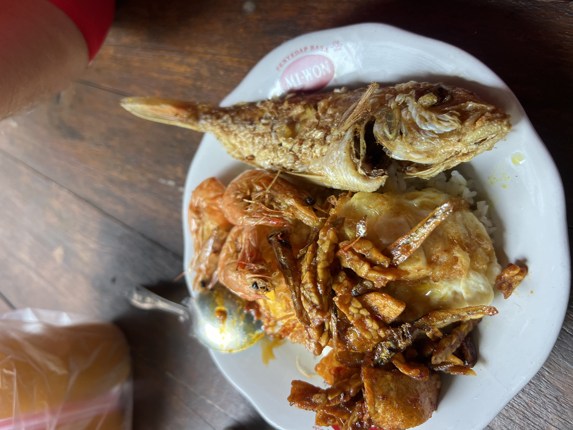 A plate of food sits on a wooden table. The plate contains rice, a fried fish, several shrimp, and a mixture of fried tempeh and small dried fish. A spoon rests on the plate, partially scooping some of the tempeh and fish. In the background, a plastic cup with a pink straw is visible. The overall scene suggests a casual meal. The location is Kuta, Indonesia, a popular tourist destination known for its beaches and restaurants.
