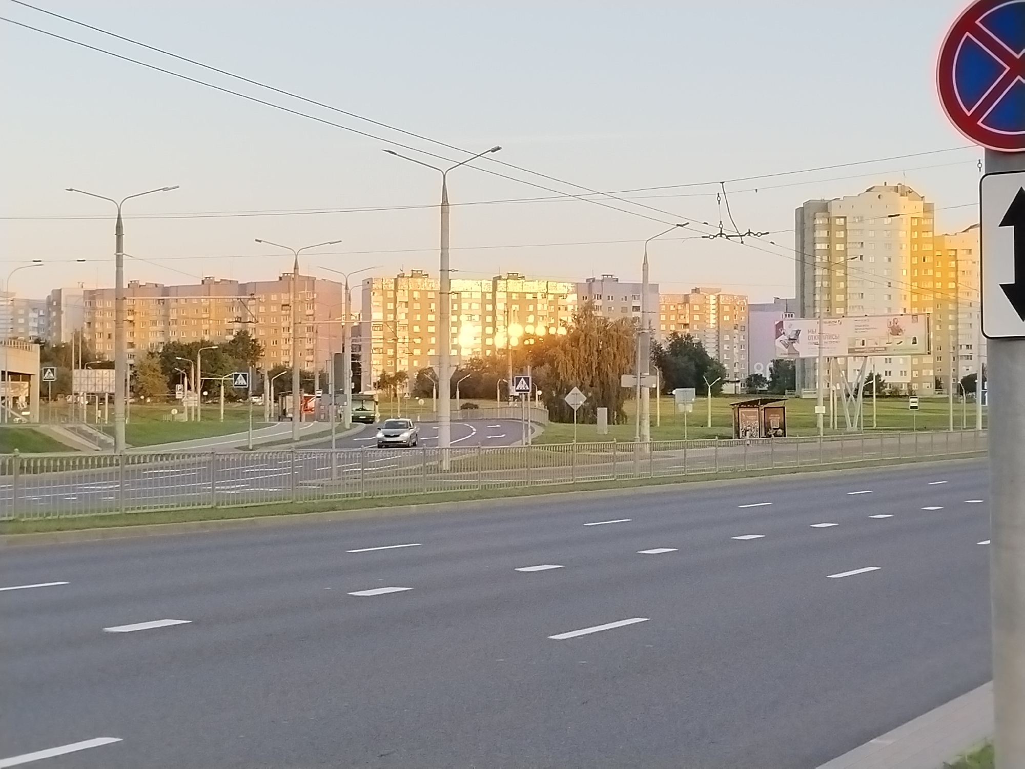 The image captures a street scene in Hrodna, Belarus, during the golden hour. In the foreground, a wide asphalt road with white lane markings stretches towards the horizon. A low metal fence runs parallel to the road, separating it from a grassy area. Beyond the fence, a multi-lane road curves, with a pedestrian crossing marked by a diamond-shaped sign with a pedestrian symbol. Several cars are visible on this road, including a silver sedan and a green vehicle.
In the middle ground, a line of apartment buildings with numerous windows rises against a pale sky. The setting sun casts a warm glow on the facades of these buildings, reflecting brightly on some sections. Streetlights with multiple lamps are positioned along the road. A small bus stop shelter with a curved roof is also visible.
In the background, more apartment buildings of varying heights and architectural styles can be seen. Overhead power lines crisscross the sky. A traffic sign with a red circle and a diagonal bar over a blue circle, indicating a prohibition, is present on the right side of the image, accompanied by a black and white directional arrow sign below it. The overall atmosphere is one of urban tranquility as the day ends.