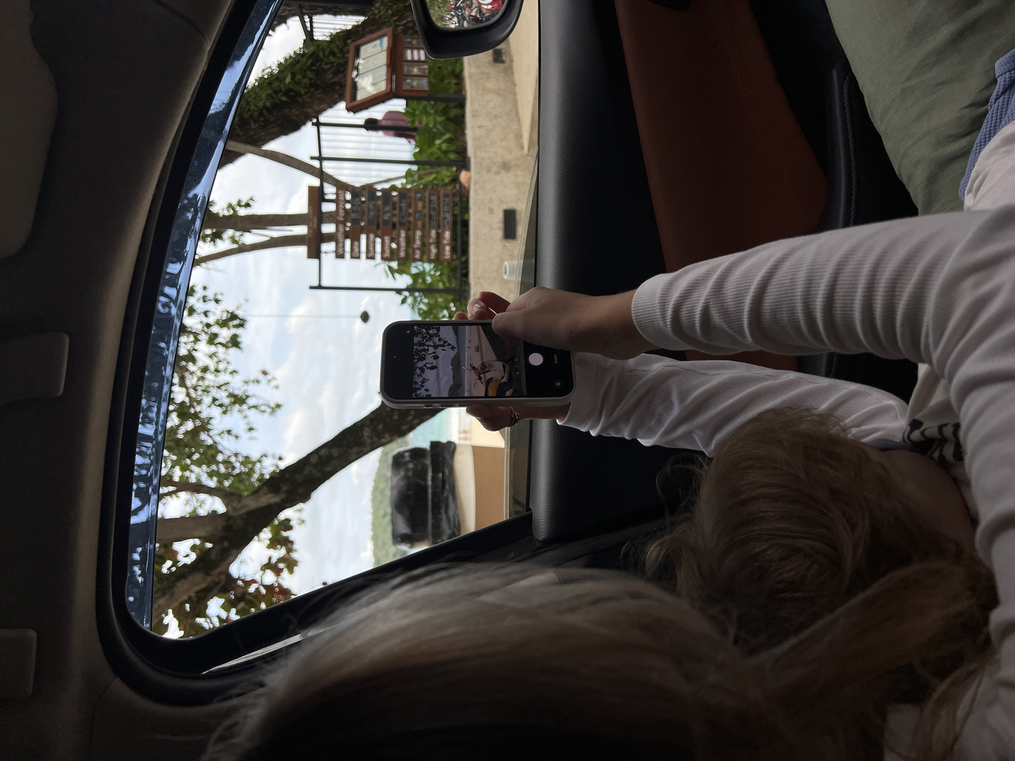 A person is in a car, taking a photo with their phone of a view outside the car window. The view appears to include a weather forecast board, trees, and possibly a beach or ocean in the distance. The person taking the photo is in the passenger seat. The location is Unknown, Unknown.