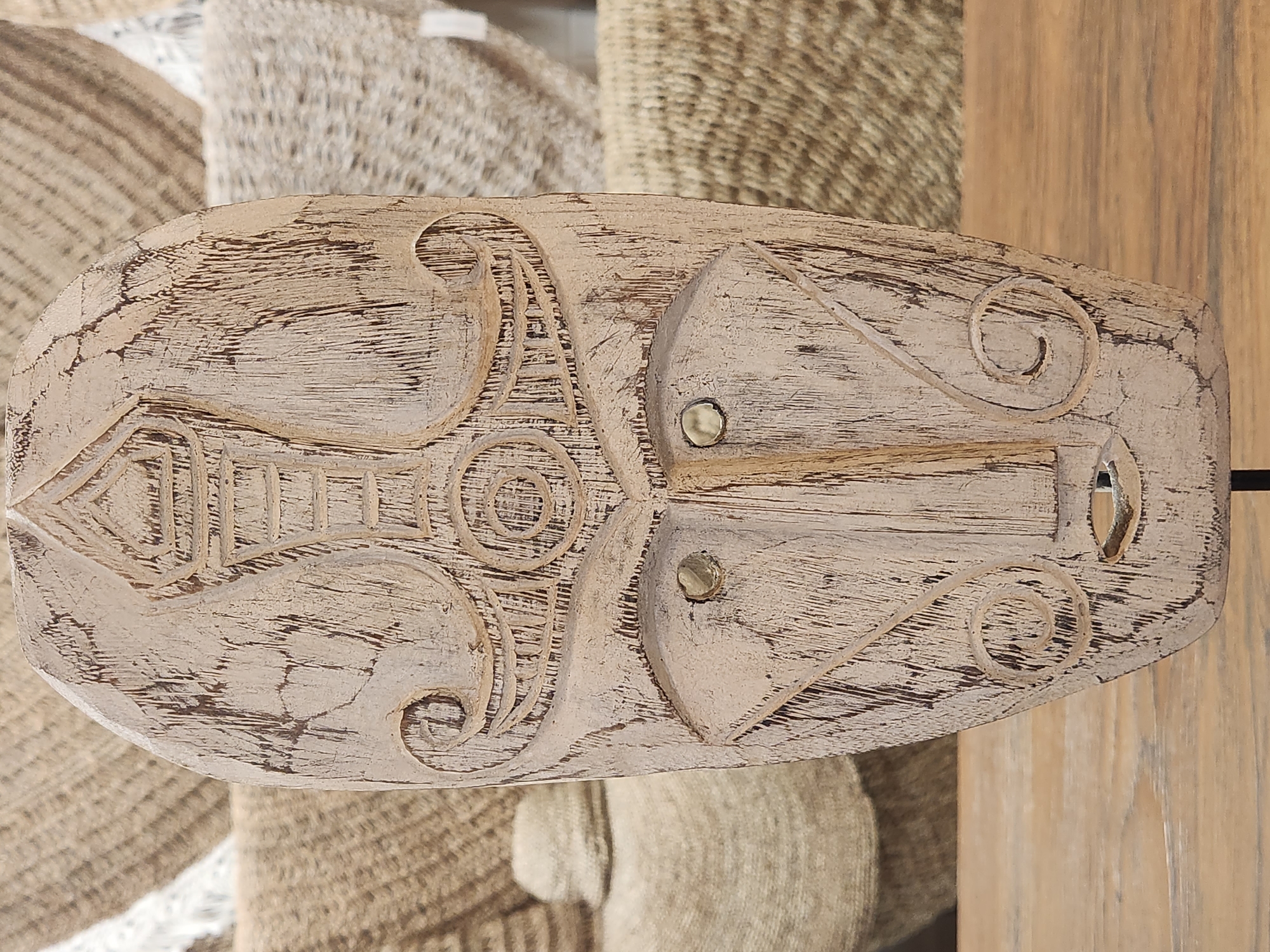An aged, carved wooden mask with an elongated oval shape and a distressed, off-white finish is displayed on a dark metal stand. The mask features a prominent nose, two circular indentations for eyes with visible metallic fasteners, and a small, carved mouth. Decorative motifs, including swirling patterns and geometric designs, are etched into the surface. The mask is positioned in the foreground, resting on a wooden surface. In the background, there is a blurred collection of woven baskets and textiles, suggesting a marketplace or shop setting. The overall lighting is soft and diffused. The location is not specified in the image or provided context.