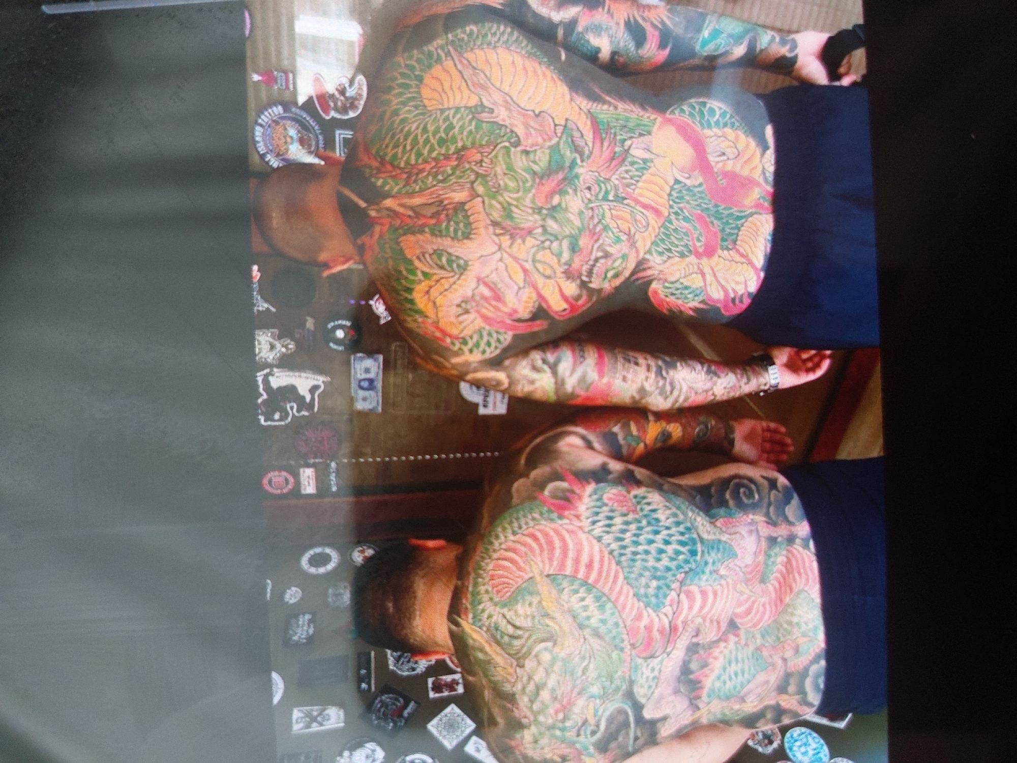 Two shirtless men wearing dark blue shorts lie side-by-side on a wooden surface in what appears to be Tsarevo, Bulgaria. Each man has an extensive tattoo of a dragon covering their entire back and arms. The tattoos feature vibrant colors including red, green, orange, and blue. A collection of stickers and small objects is scattered on the wooden surface next to the men.