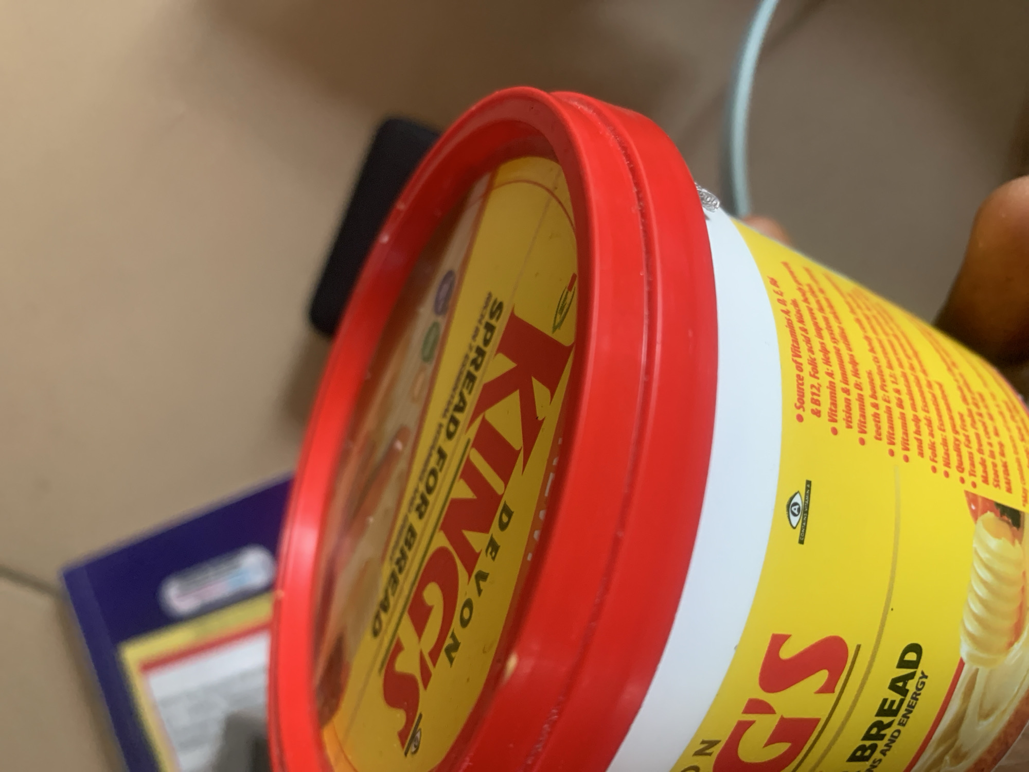 The image is a close-up, low-angle, eye-level shot of a yellow tub of "Devon Kings Spread for Bread" with a red lid. The tub is partially visible, with the brand name "KINGS" prominently displayed in large, red, block letters. Below this, "SPREAD FOR BREAD" is written in smaller red text. The tub has a slightly reflective surface. To the left of the brand name, the word "DEVON" is visible in smaller red text.
The tub is positioned on a light-colored surface, possibly a table or countertop. The background is blurred and out of focus, showing hints of other objects, including what appears to be a blue and yellow rectangular item, possibly packaging, to the left of the tub. A dark, elongated object, possibly a phone or electronic device, is also visible in the background behind the tub. The lighting is natural and appears to be coming from above and to the right, casting subtle shadows. The overall impression is that of a product shot or a casual photograph taken in an indoor setting. The location in Jauro Yinu, Nigeria, is not discernible from the visual information in the image.