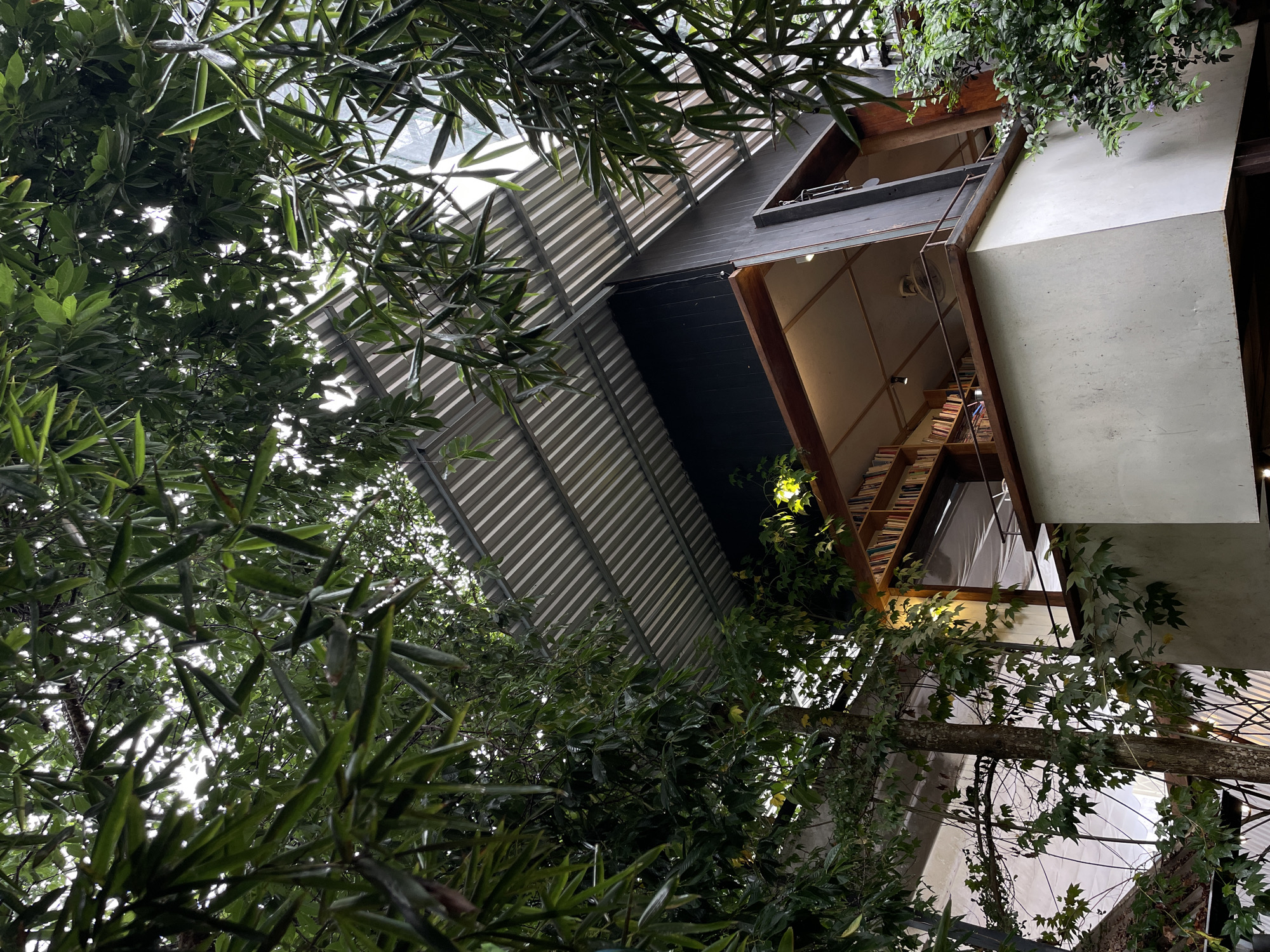 Looking up, a modern building is partially obscured by lush green foliage. The structure features a gray corrugated metal roof, a dark blue section, and wooden accents. An open area with shelves filled with books is visible. The building is constructed around and incorporates trees. The sky is partially visible through the foliage. This scene is likely located in Lâm Viên - Đà Lạt, Vietnam.