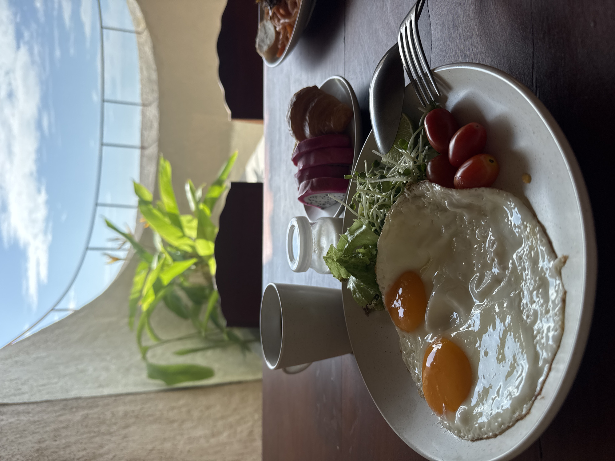This image captures a serene breakfast or brunch setting on a polished dark brown wooden table, viewed from a slightly elevated, overhead perspective.
The central focus is a light grey speckled plate holding two perfectly fried eggs with vibrant orange yolks and crispy whites, accompanied by a fresh green salad of lettuce and sprouts, and four bright red cherry tomatoes. A silver fork and spoon rest on the plate's rim.
To the left, a smaller oval dish contains several slices of visually striking pink and white dragon fruit, paired with a golden-brown croissant. Partially visible in the top background is another bowl with a savory dish, possibly a stew or stir-fry. A plain light grey mug and a small clear condiment bottle are also present on the table.
The background features a large, arch-shaped window, allowing abundant natural light to flood the scene. Through the window, a clear blue sky with white clouds is visible, alongside green foliage from a plant, suggesting an outdoor connection. The scene evokes a peaceful, healthful meal enjoyed during the day, possibly in a pleasant, resort-like environment in a tropical location such as Bac Nha Trang Ward, Vietnam. No text is visible in the image.