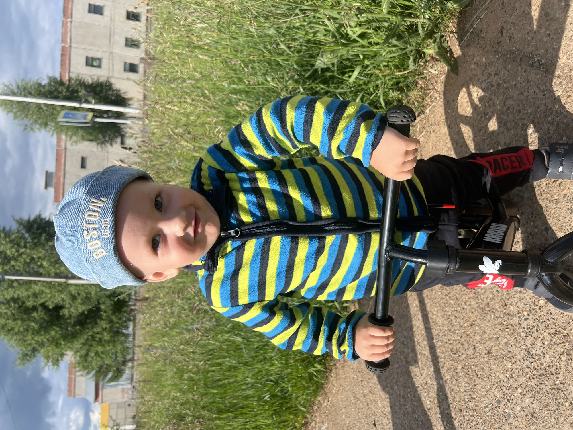 A young child wearing a light blue beanie with "BOSTON 1630" embroidered on it and a striped fleece jacket in blue, yellow, and black, is positioned outdoors. The child is holding the handlebars of a black balance bike. The background features tall green grass, a paved pathway, and a building with multiple windows. The sky is partly cloudy. The setting appears to be an outdoor urban environment, potentially in Markovsky, Russia, given the presence of the building and the general appearance of the surroundings.