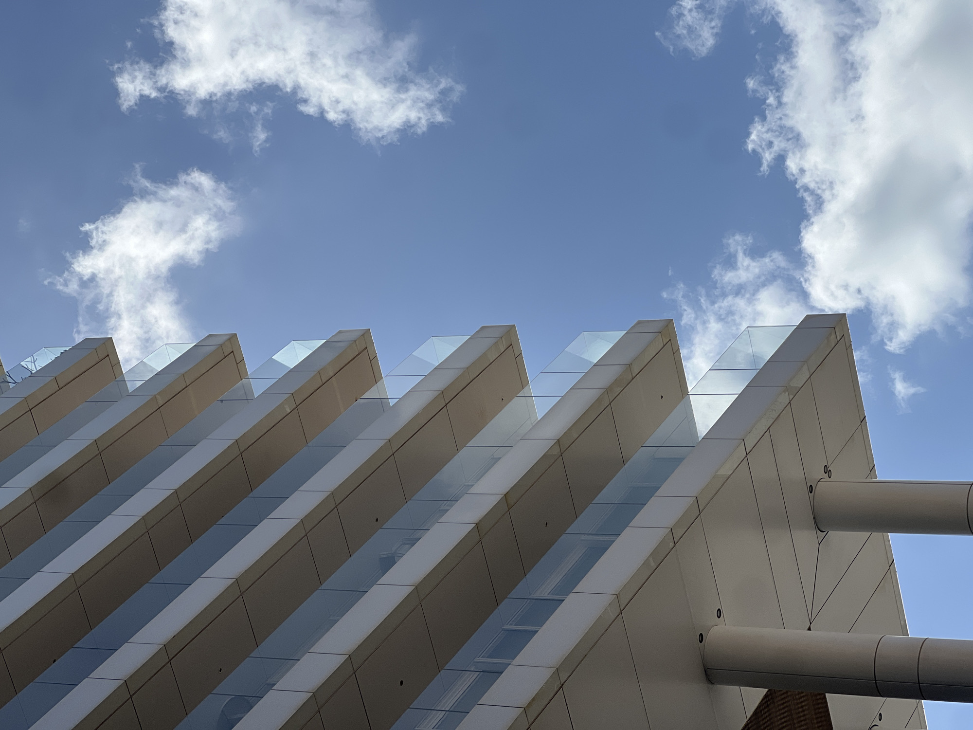 A low-angle shot reveals a modern building with a distinct, repeating geometric design against a backdrop of a blue sky scattered with fluffy white clouds. The building's facade is characterized by a series of angular, white panels that create a rhythmic pattern. Glazing or clear panels are set between these white sections, allowing light to penetrate the interior. Cylindrical elements, possibly support structures, are visible on the right side of the frame, attached to the building's facade. The sky above is clear and blue, with a few scattered cumulus clouds, adding depth and brightness to the image. This building is located in Tel-Aviv, Israel.