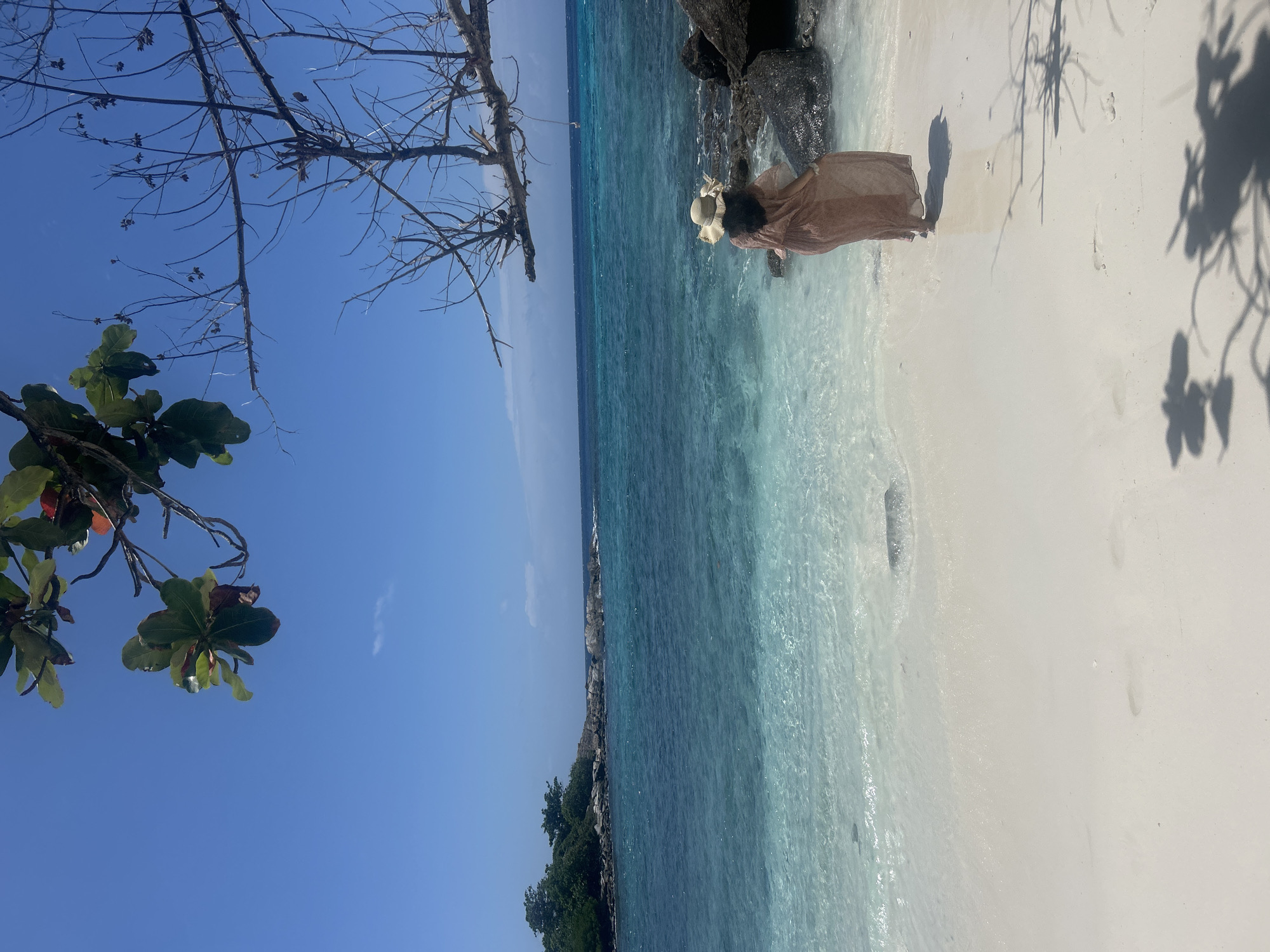 A person stands on a white sand beach in Rawai, Thailand, facing the clear blue ocean. The person is wearing a wide-brimmed hat and a flowing, translucent tan dress. The water is calm and appears shallow near the shore, with visible sand and rocks beneath the surface. In the distance, a rocky island covered in green trees is visible. The sky is a vibrant blue, with a few bare branches from a tree extending into the foreground from the top of the frame. Shadows of the tree branches are cast on the sand. The scene depicts a serene tropical environment.