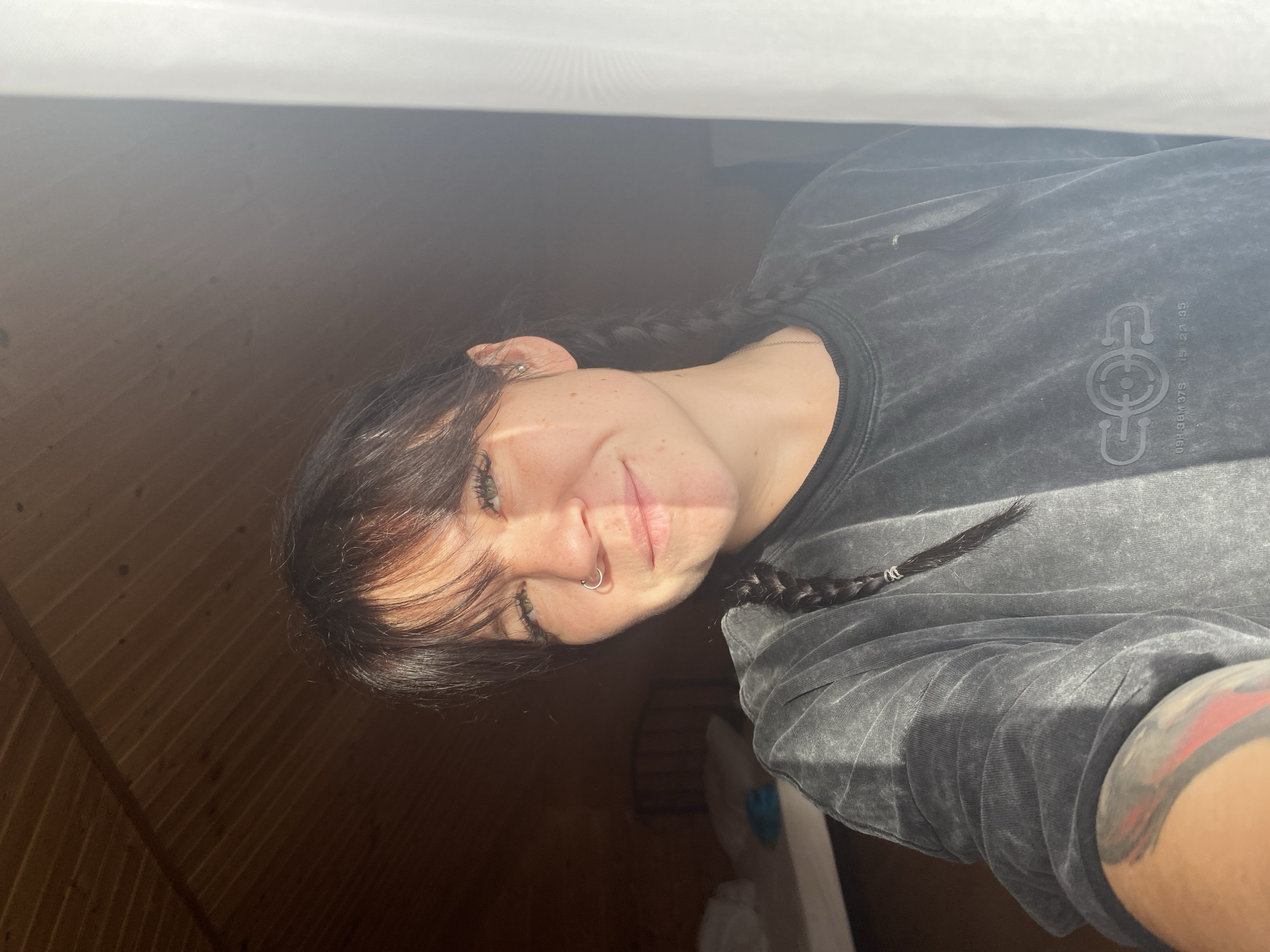 The image shows a young woman with dark hair styled in two braids, wearing a dark grey, acid-washed sweatshirt. She has a nose ring and a piercing in her left ear. Her face is partially illuminated by sunlight, creating a shadow across the right side of her face. She is smiling and looking towards the camera. Behind her, a wooden ceiling is visible, and to the right, a white curtain is present. In the blurred background to the left, a bed frame can be seen. The overall setting appears to be an indoor space, possibly a room with a sloped ceiling. The image does not contain any contextual information that would link it to Krasnoflotskoe, Russia.