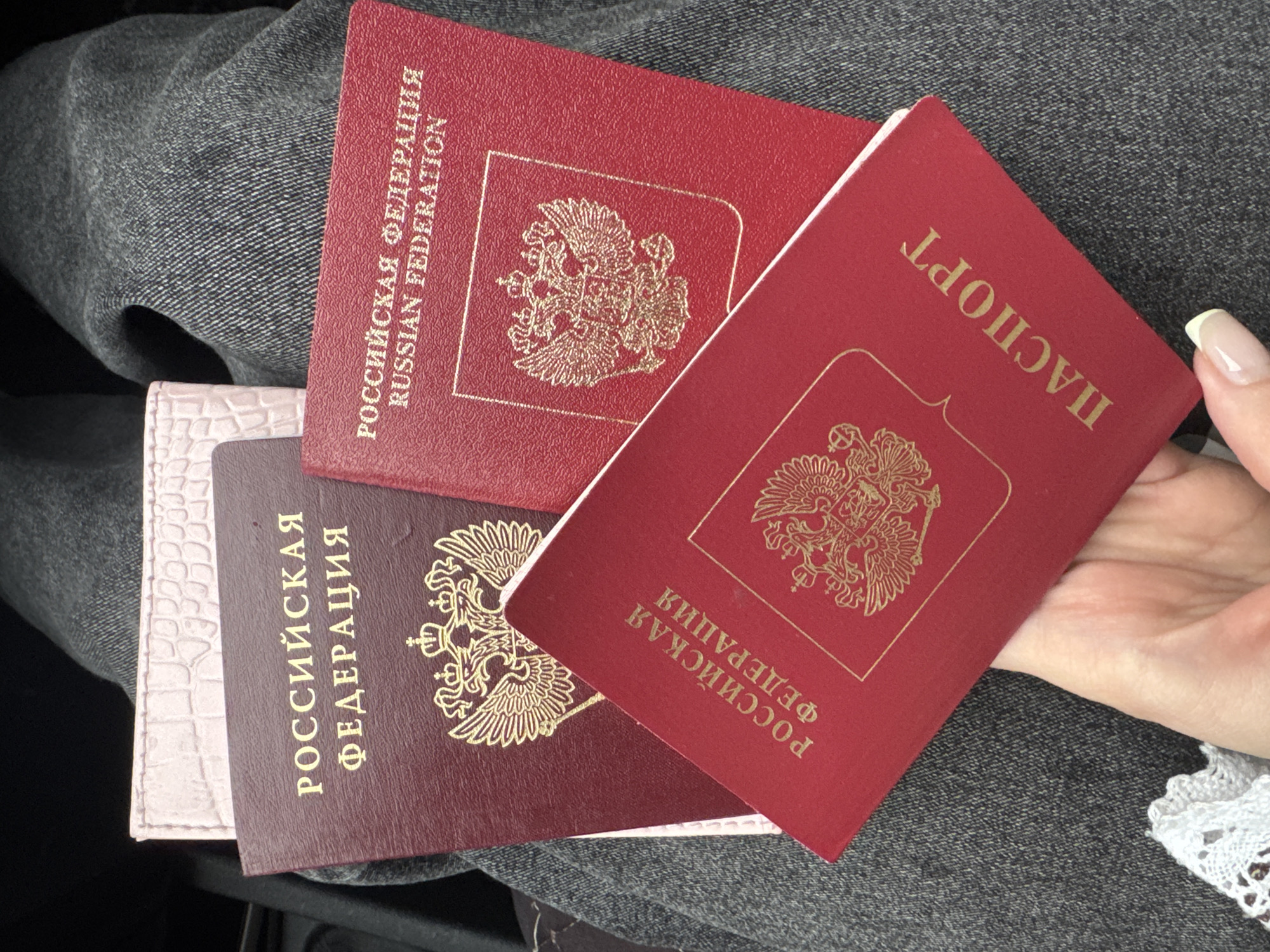 A person's hand, with painted fingernails, holds two red Russian passports. The passports are layered on top of a dark grey denim-like fabric. One passport is titled "РОССИЙСКАЯ ФЕДЕРАЦИЯ" (Russian Federation) in gold lettering and features a golden double-headed eagle emblem. Another passport, partially visible beneath the first, also displays "РОССИЙСКАЯ ФЕДЕРАЦИЯ" and a similar golden emblem. A third, lighter pink, textured document peeks out from behind the passports. The scene appears to be indoors or in a shaded area, given the lighting. There are no discernible city landmarks or specific geographical markers indicating a location in Perm, Russia, or any other city.