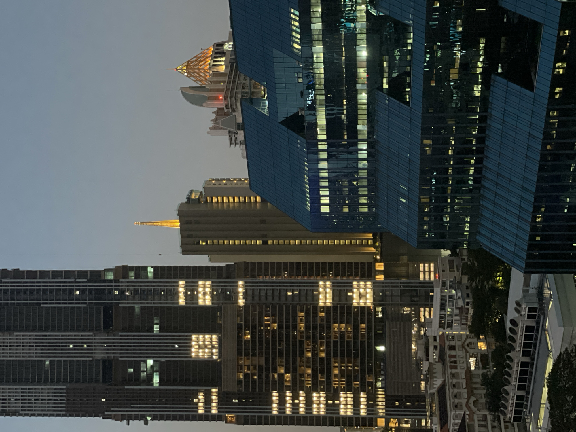 The image depicts a nighttime cityscape in Bangkok, Thailand, featuring several illuminated skyscrapers. The buildings showcase a variety of architectural styles, with one structure displaying a distinctive angular design in shades of blue, and others exhibiting grid-like patterns created by numerous lit windows. In the background, a structure with a golden spire can be observed. The sky appears overcast, contributing to the overall mood of the scene. No people are visible in the shot.