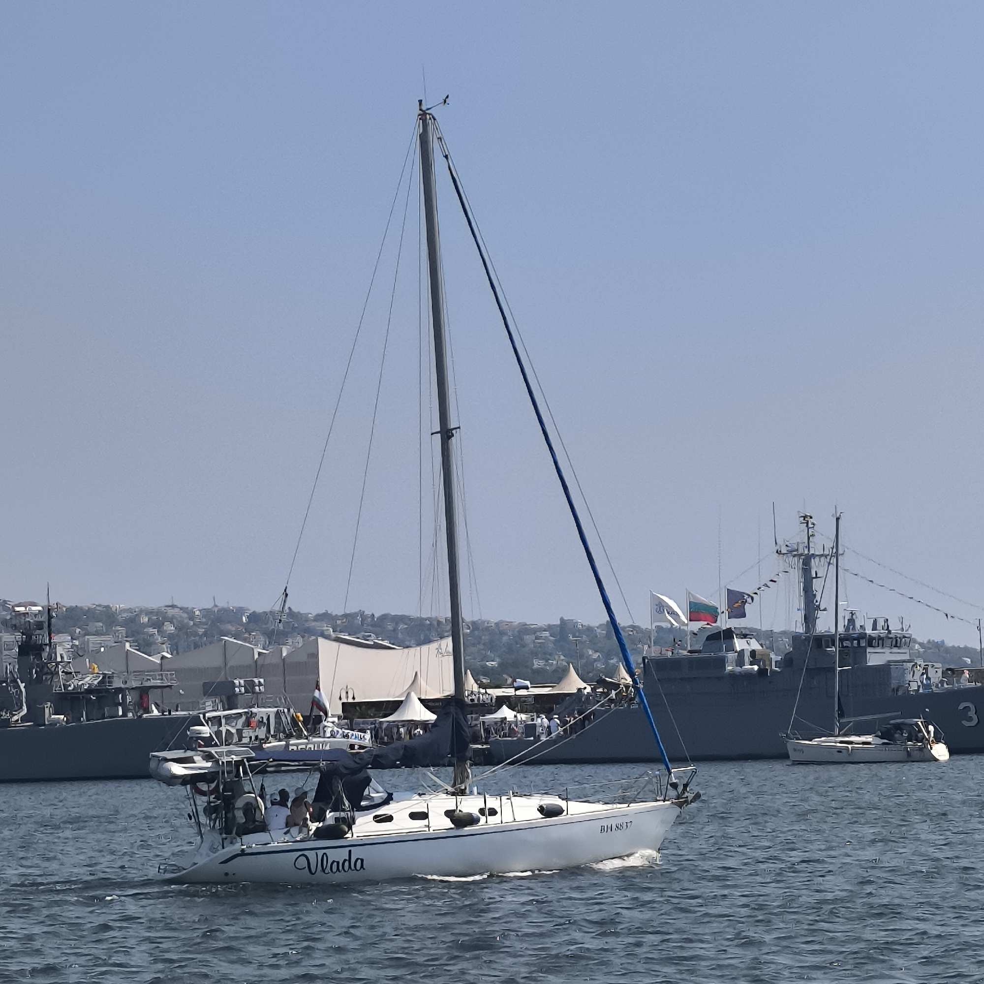 A daytime, wide shot captures a sailboat gliding across rippling blue water in the foreground, with a large, grey naval vessel anchored in the background. The sailboat, named "Vlada," has several passengers on board. Behind the naval ship, a city skyline is visible, with buildings nestled on a hillside. The scene appears to be set in the Port of Varna, Bulgaria, likely during an event or celebration, given the presence of flags and what looks like a festive gathering onshore. The sky is clear and blue.