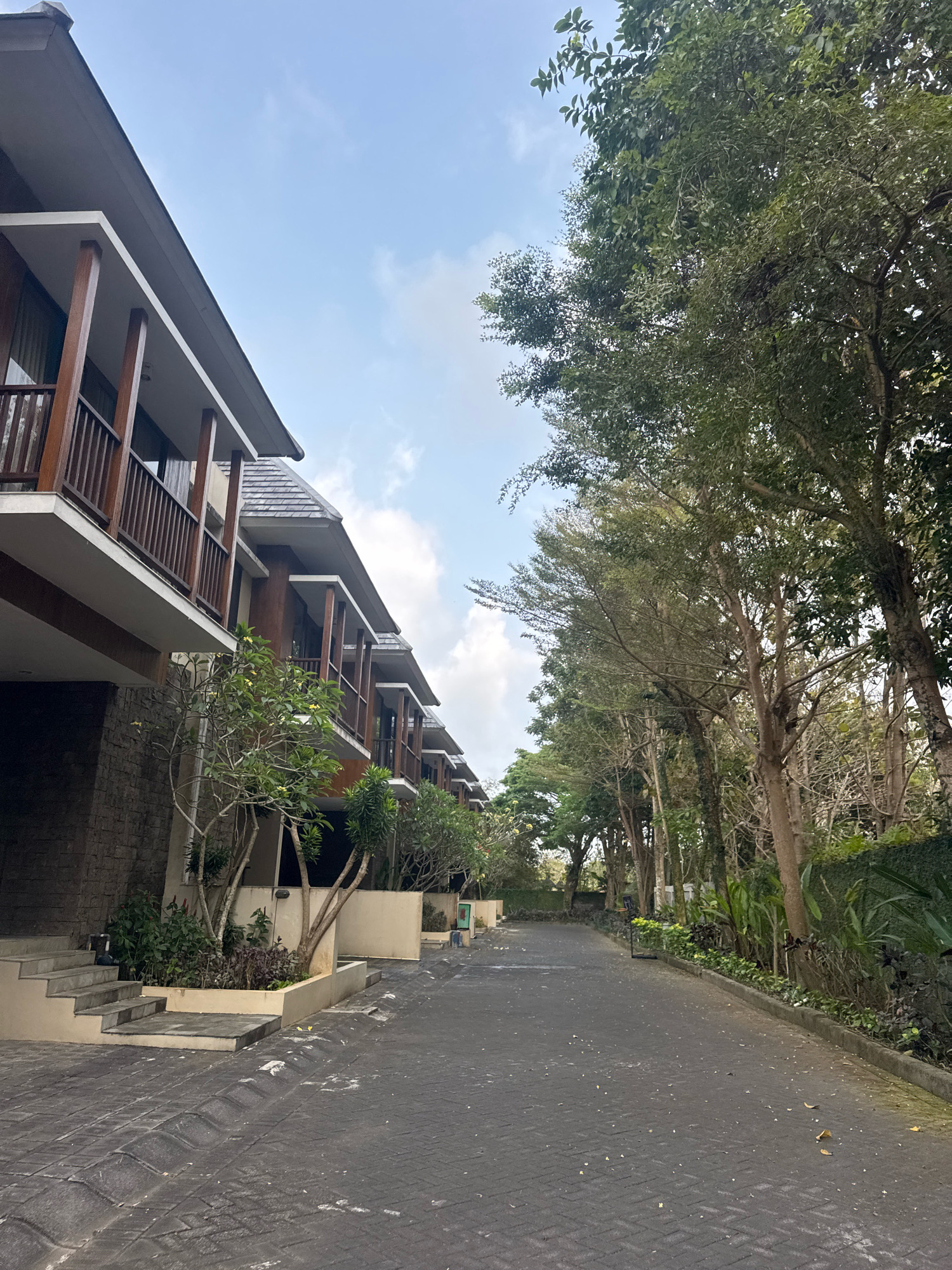 The image shows a paved pathway in Kuta Selatan, Indonesia, bordered on the left by a row of modern multi-story buildings with wooden balconies and on the right by a dense line of trees. The pathway is constructed of dark interlocking bricks, with a slight incline visible. The buildings feature stone bases and upper levels with balconies supported by wooden pillars. Small trees and shrubs are planted in raised planters along the building's edge. The trees on the right are lush and green, creating a natural canopy over the pathway. The sky above is mostly clear with a few scattered clouds. No people are visible in the scene.