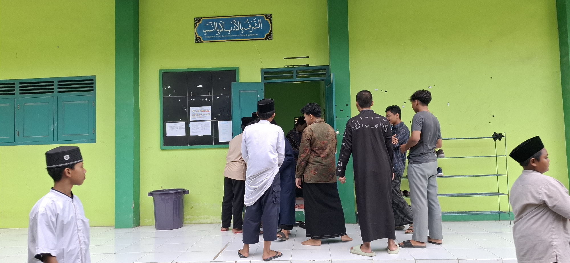 In the city of Wonorejo, Indonesia, a group of boys and young men, some wearing traditional Islamic attire such as peci caps and long robes, are gathered outside a building with a bright green facade. The building has two prominent teal-colored windows with louvers and a door to the right. Above the door, a blue and white sign with Arabic calligraphy is mounted on the wall. To the left of the door, a bulletin board with papers is visible. A purple trash bin stands near the wall. In the background, a metal shoe rack is present. The scene appears to be an outdoor area of a school or religious institution, possibly for morning assembly or a break period.