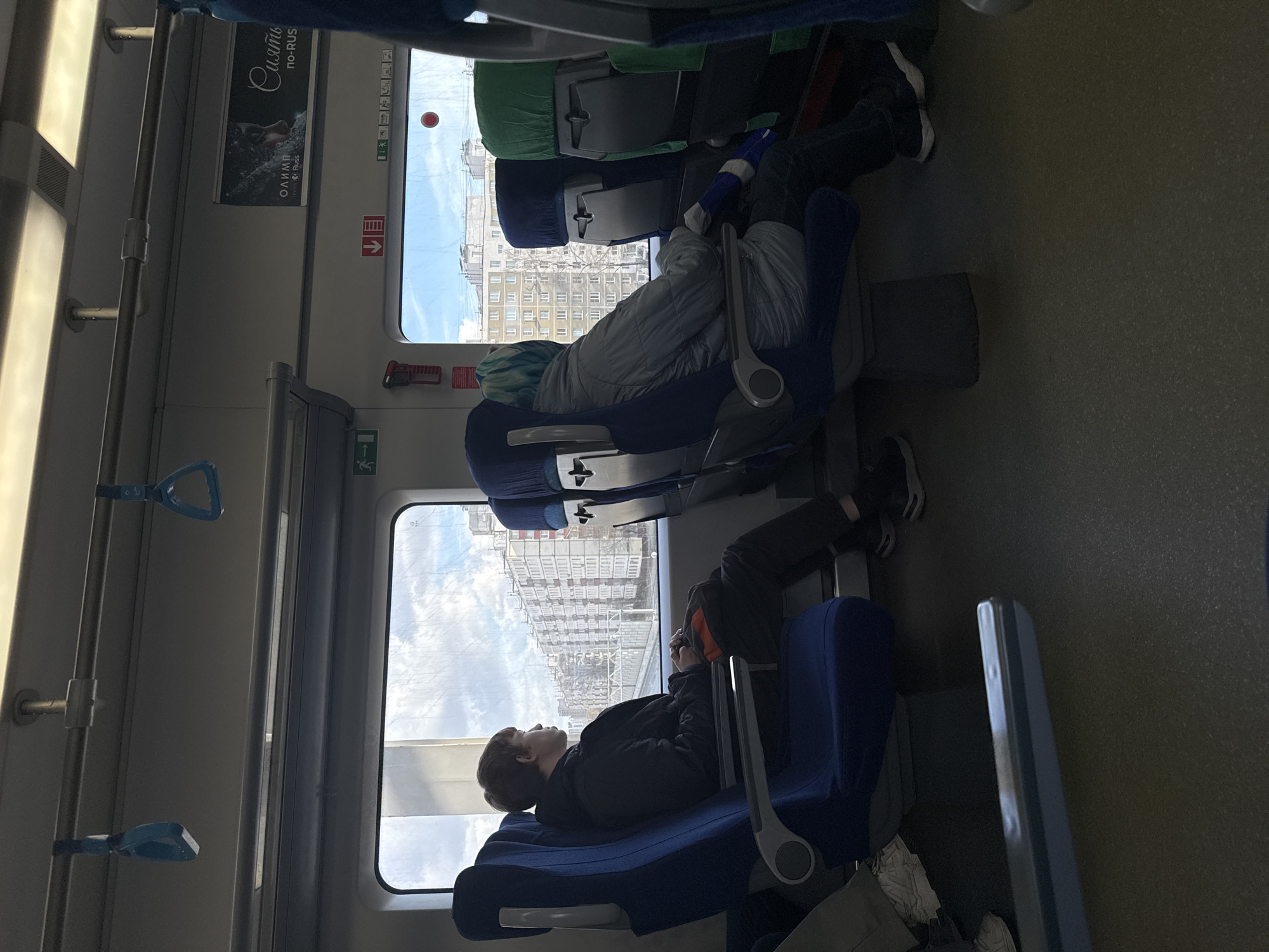 The interior of a train carriage is depicted. Two individuals are present. One person with dyed blue hair is seated and wearing a grey jacket. Another person is reclining, dressed in dark clothing. Outside the window are buildings indicating that the location is the city of Khimki, Russia. The scene appears to be taken during daytime. The train car is fairly modern with blue seats.