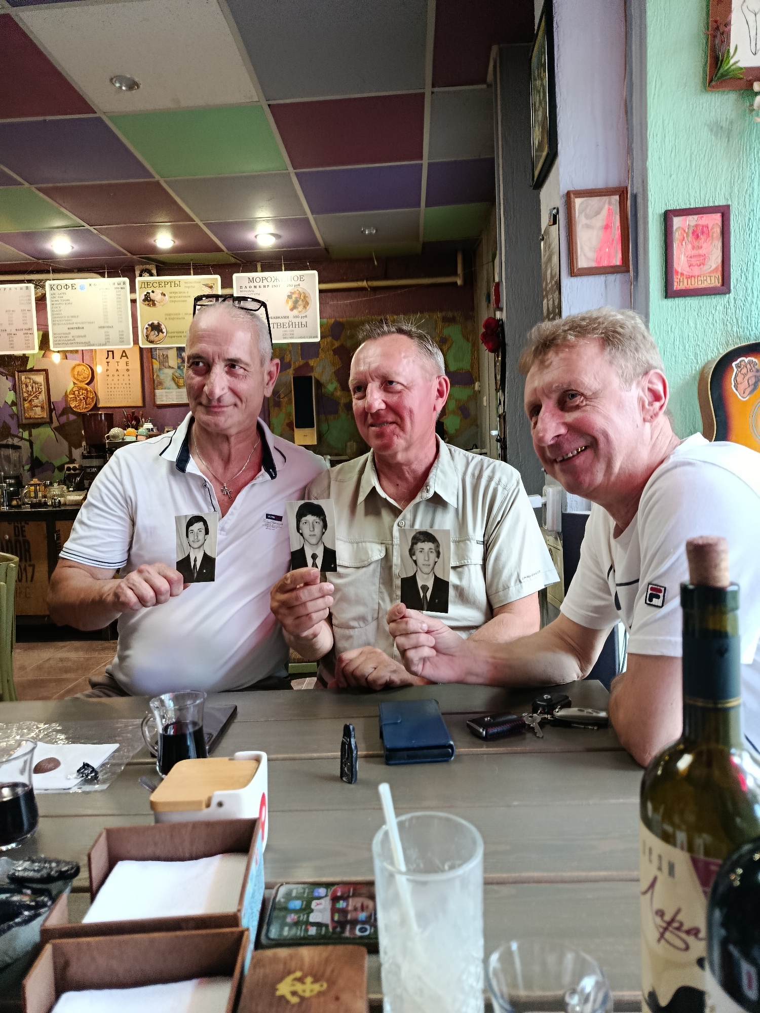 Three middle-aged Caucasian men are seated at a wooden table, likely in a cafe or restaurant. The man on the left is wearing a white polo shirt and holding a small black-and-white photograph of a young man in a suit. The man in the middle is wearing a light-colored button-down shirt and also holding a similar photograph of a young man in a suit. The man on the right is wearing a white t-shirt with a red and white logo and appears to be smiling. In front of the men are drinks in glasses, a bottle of wine, a small white container with a wooden lid, and other miscellaneous items. The background shows a colorful ceiling, posters on the wall, and a guitar. The overall impression is a casual gathering. The presence of Cyrillic script on posters and potentially on the wine bottle suggests the location is Russia, and the sign on the wall that reads "САРАТОВ" confirms the city is Saratov.