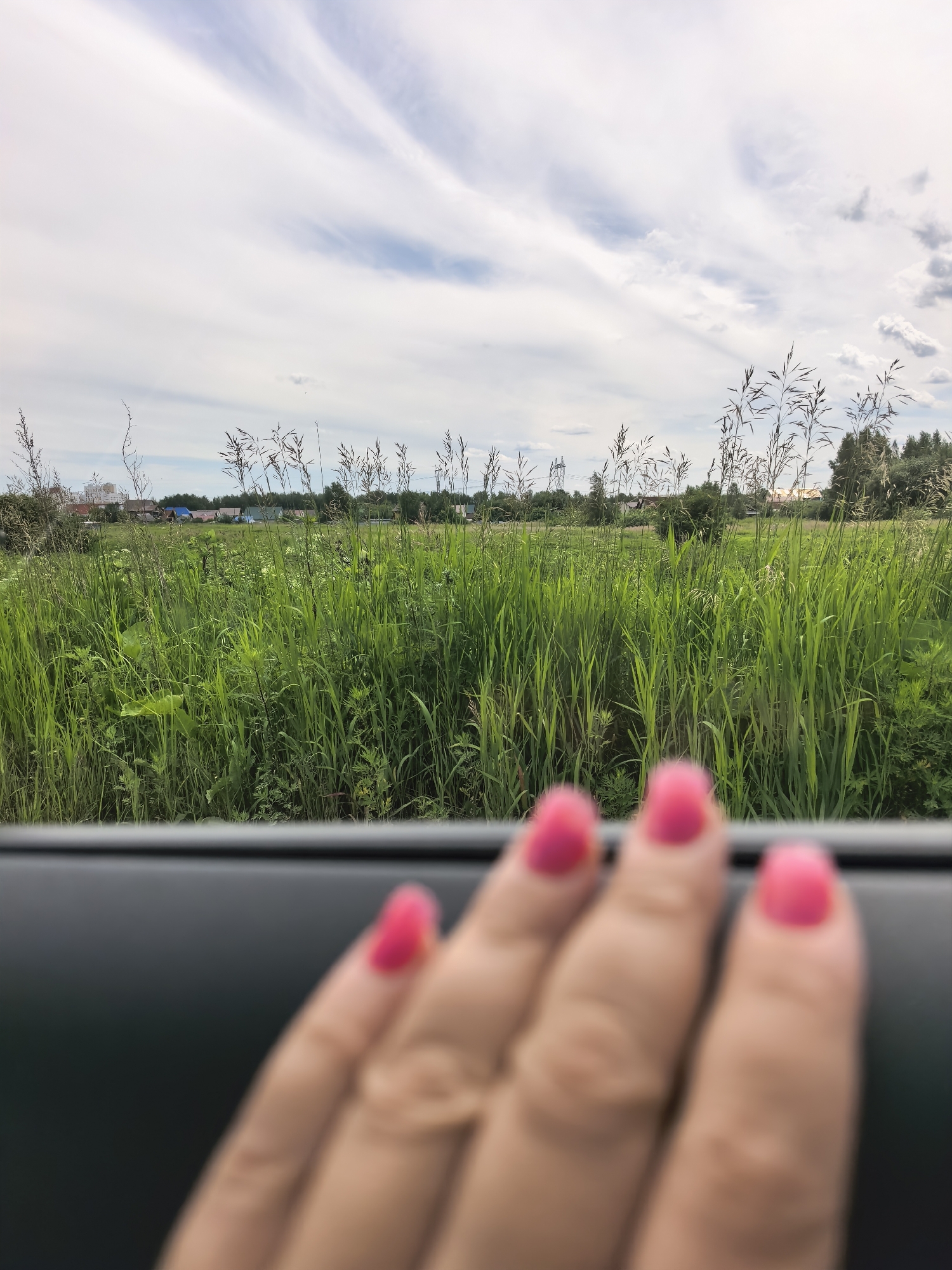 An outdoor, medium-shot photograph captures a hand with bright pink fingernails resting on a dark surface, likely the interior of a vehicle. The hand is out of focus, with the pink nails being the clearest elements. Beyond the hand, a lush, green field extends into the distance, dotted with tall grasses and wildflowers. In the background, a line of trees and several buildings, including houses with colorful roofs, are visible. The sky above is a mix of white and pale blue clouds, with wispy formations stretching across the frame. The scene suggests a rural or suburban setting, possibly a scenic drive through the countryside.