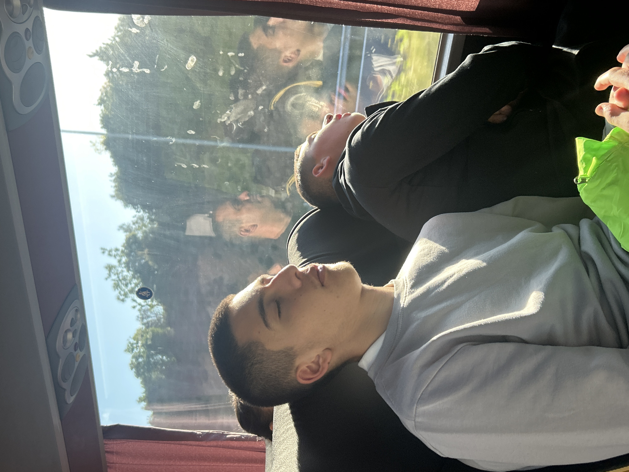 Two young men are sleeping on a bus. The man in the foreground is wearing a light gray sweater and has short, dark hair. He is leaning against the seat. Behind him, a man in a dark hoodie is also sleeping with his arms crossed. The reflection of the men in the bus window shows the trees outside. The bus window has a sticker that shows the handicapped seat symbol. The picture was taken during the daytime with a lot of sunlight coming through the window. The location is likely the Municipal Unit of Kato Olympos, Greece.