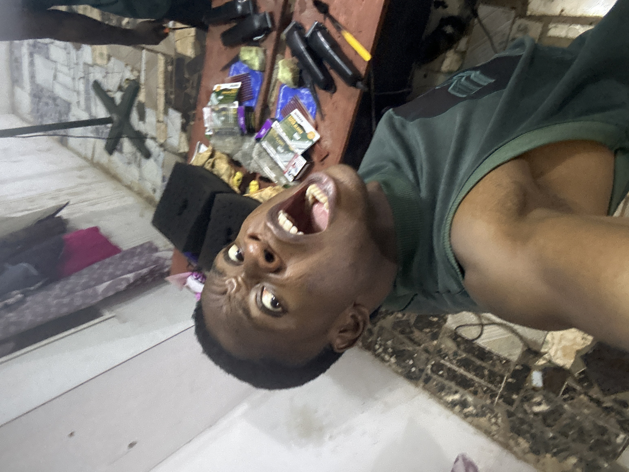 A young Black man is prominently featured in this image, rotated 90 degrees clockwise, looking directly into the camera with a wide-open mouth, suggesting a shout, laugh, or exaggerated expression. He wears a dark green t-shirt. His eyes are wide, and his teeth are clearly visible.
The scene appears to be an indoor setting, possibly a barber shop or a personal grooming area within Ikorodu, Nigeria. To his right, a wooden surface serves as a table or counter, laden with grooming tools. These include multiple hair clippers, blue plastic combs, and brushes, one with a distinct yellow handle. Several small product packets or boxes are scattered among the tools, with the brand name "WEHAAO" clearly legible on one. Brown or gold-colored wrappers are also visible on the table.
In the background, a light-colored wall shows signs of wear and features a prominent, dark "X" painted or drawn on it. A dark, thin pole-like object extends from the left side of the frame towards the "X." The floor is tiled in a light color and shows some debris. A small pink fabric item is on the floor in the lower left. In the upper left background, another person's arm or shoulder, wearing a dark top, is partially visible. The overall impression is one of a functional, somewhat cluttered space.