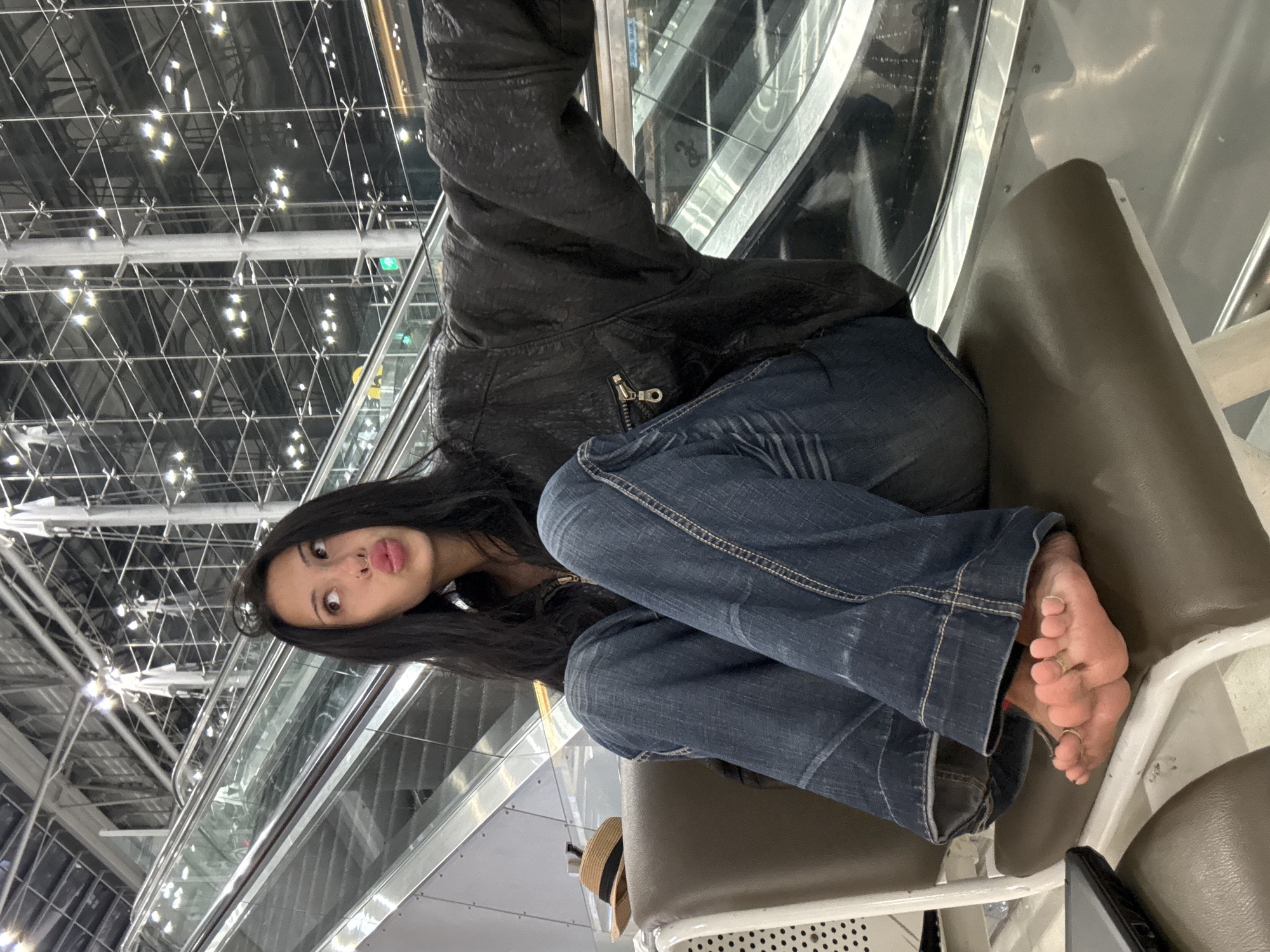 A young woman with long, dark hair sits on a brown chair in what appears to be an airport terminal. She is wearing a black leather jacket and blue jeans, with her bare feet resting on her lap. Her toes are adorned with rings. The background features escalators and a modern, industrial-style ceiling with numerous lights. To her left, a straw hat with a black ribbon sits on an adjacent chair. The overall scene is well-lit, suggesting an indoor public space.