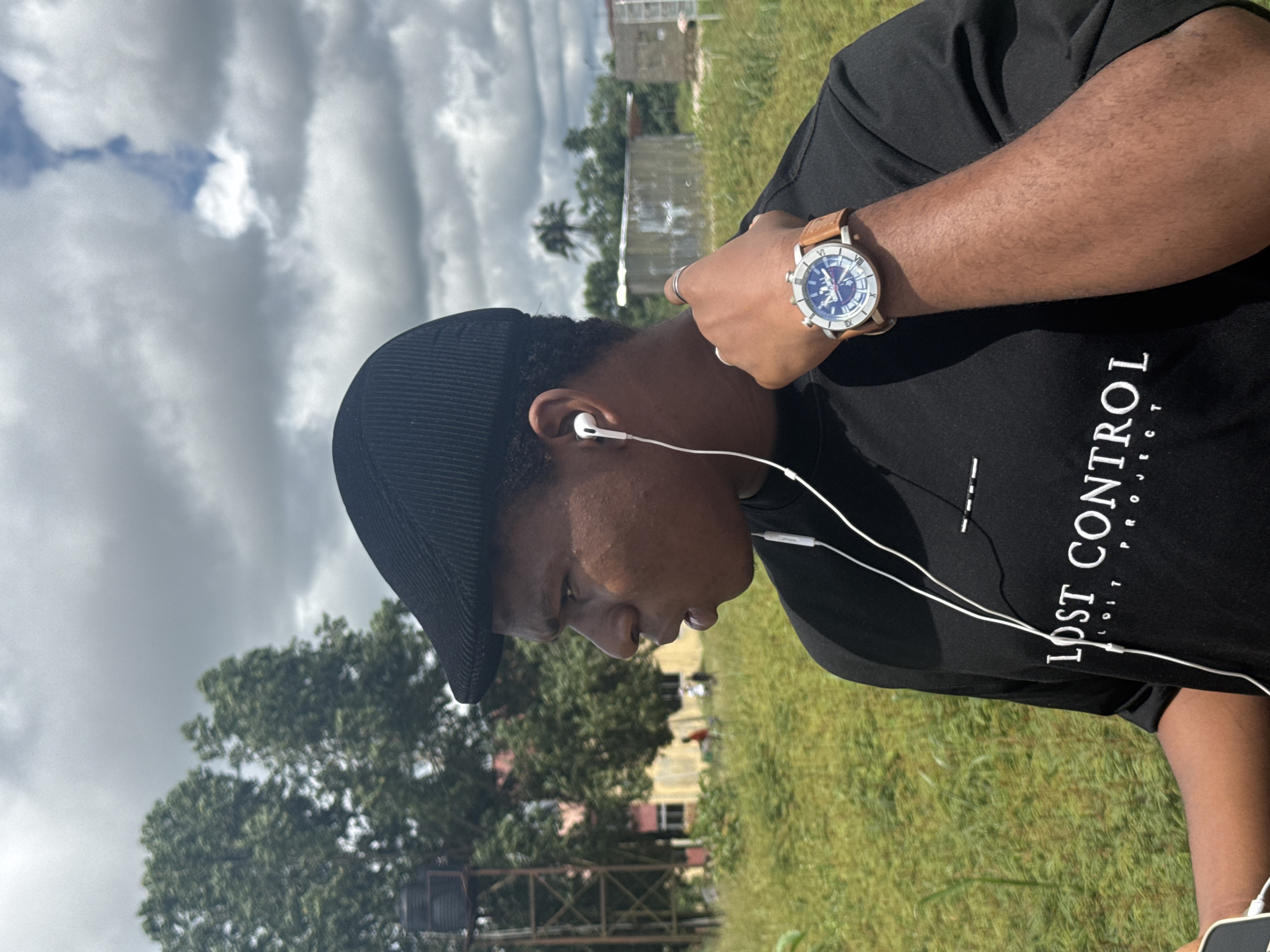 A young Black male wearing a black cap, black t-shirt with white text that reads "LOST CONTROL", and white wired earbuds is shown in a medium shot outdoors. He is looking to his left with his mouth slightly open. He is wearing a watch with a brown leather strap and a blue and silver face on his left wrist. The background consists of tall green grass, trees, a water tower on the left, and some buildings and sheds further in the distance. The sky is cloudy with patches of blue. This scene is located in Ihiagwa, Nigeria.
