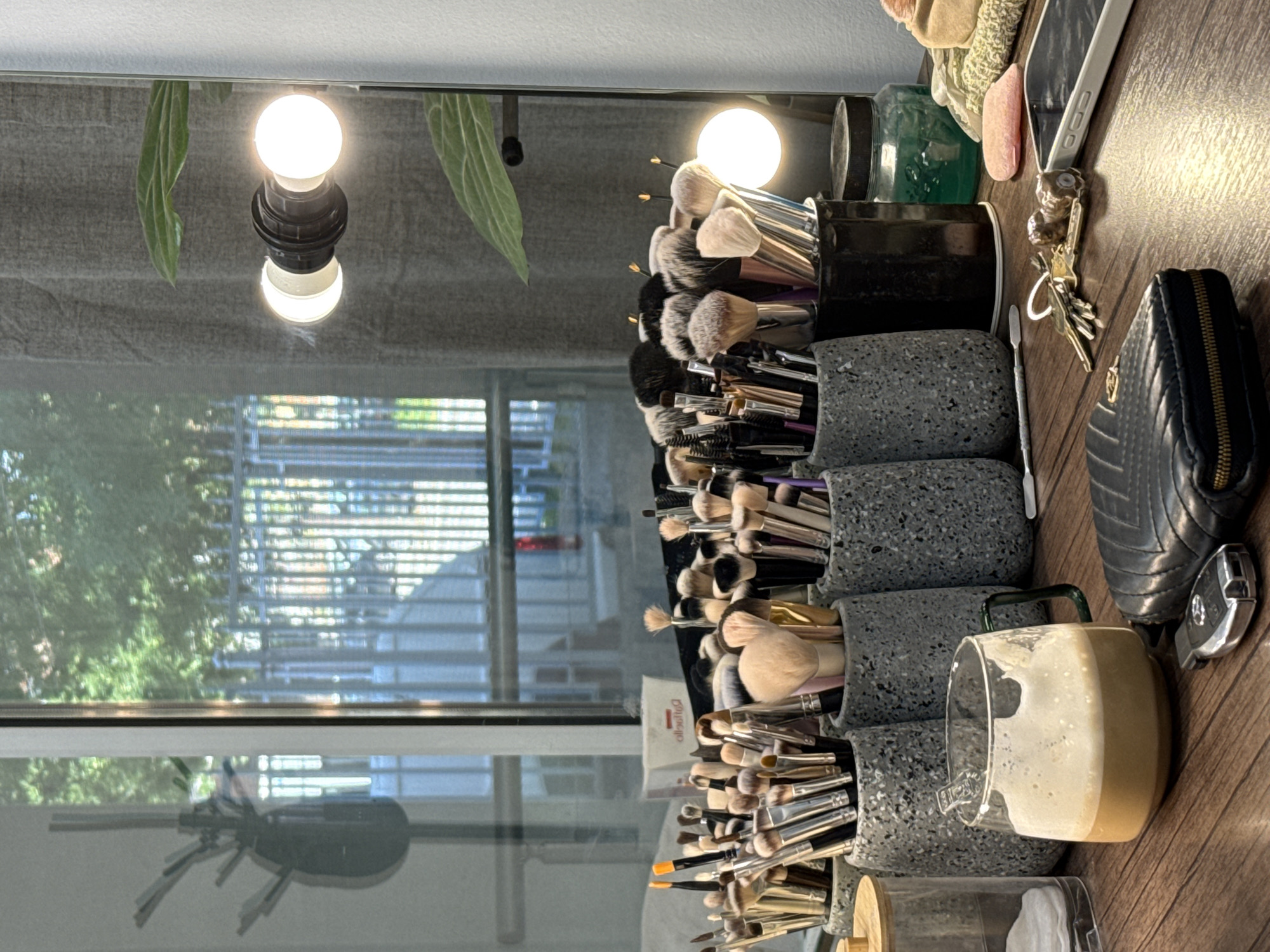 The image depicts a vanity or makeup table with a collection of makeup brushes organized in gray speckled containers. The containers are stacked and filled with various sizes and types of brushes. Adjacent to the brushes are personal items, including keys, a black wallet, and a half-empty glass of a creamy liquid, potentially a beverage. A phone and cosmetic containers are also present. The table surface is wooden, reflecting the light. In the background, a vanity mirror reflects the surroundings. The reflection reveals a window with blinds and an exterior view of greenery. Two vanity lights are visible above the makeup brushes. While the image provides no information to establish that the picture was taken in Pleven, Bulgaria.