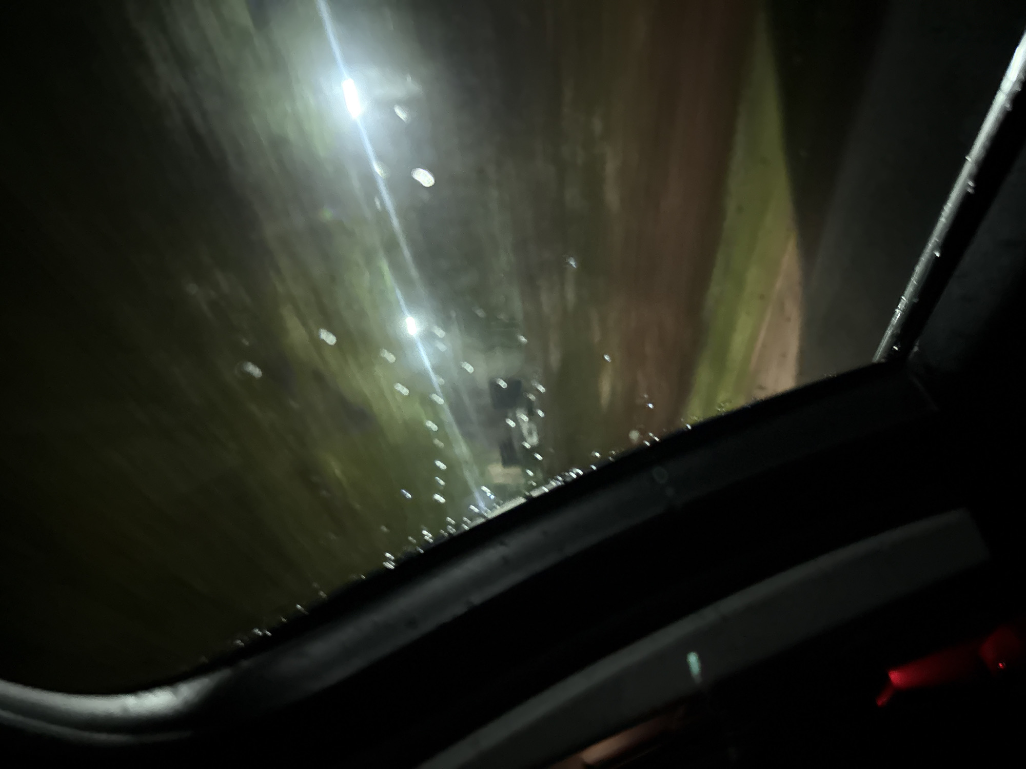The image shows a view through a wet sunroof at night. Raindrops are visible on the glass. Outside, there are blurred lights and a dark background, suggesting an urban or roadside scene. The interior of the vehicle is dark, with parts of the sunroof mechanism and frame visible. There is no indication of a specific location in Slavyani, Bulgaria or any people.