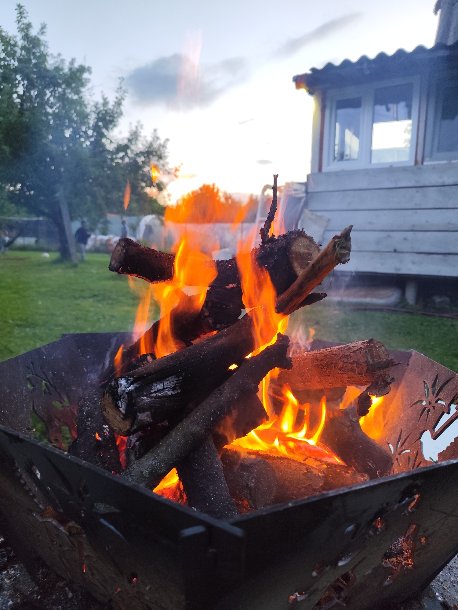 A fire burns in a decorative metal fire pit in a backyard setting. The fire pit is filled with logs that are actively burning, with flames reaching upwards. The fire pit has cut-out designs of birds and plants around its edges.
In the background, a wooden structure with white-trimmed windows and a corrugated roof is visible. The area around the fire pit is grassy, suggesting an outdoor lawn. Beyond the lawn, trees are present, and a few people are seen in the distance, one of whom appears to be kicking a soccer ball. The sky above is cloudy with hints of an evening sunset, casting a soft light over the scene. The overall mood is relaxed and reminiscent of a casual outdoor gathering. The time of day is likely dusk. The location is specified as Kolpakovo, Russia. There is no visible text in the image.