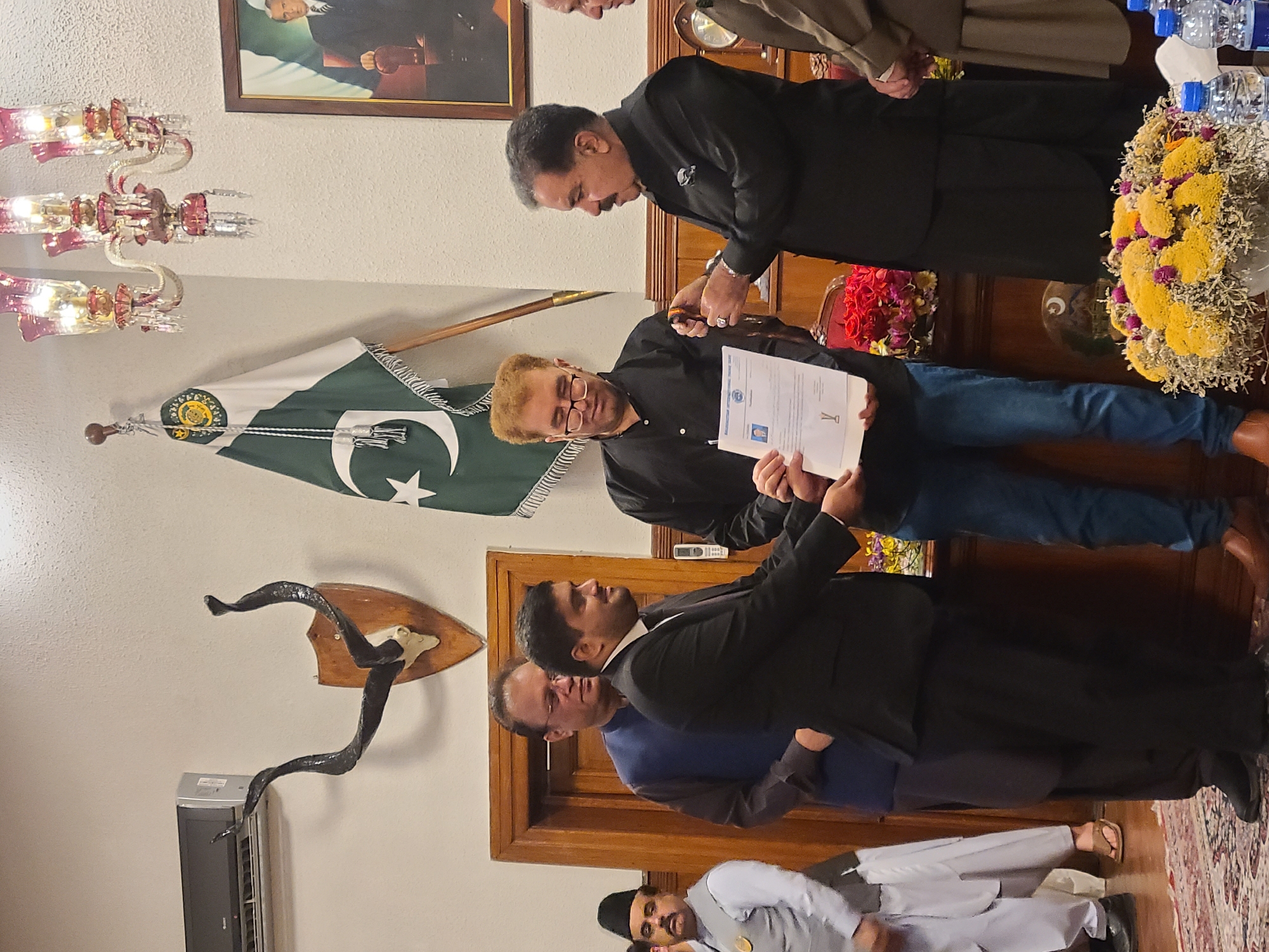 The image shows a group of men indoors. A man in a black suit is handing a certificate to another man with reddish-blond hair wearing a black shirt and blue jeans. Behind them is a flag of Pakistan. In the background, there's a framed portrait, what appears to be a chandelier, and a wooden cabinet with flowers and water bottles on top. On the left side of the photo, there's an antler mount on the wall, and what looks like an air conditioner near the floor. The men are in what is very likely a government building in the city of Quetta City Tehsil, Pakistan.