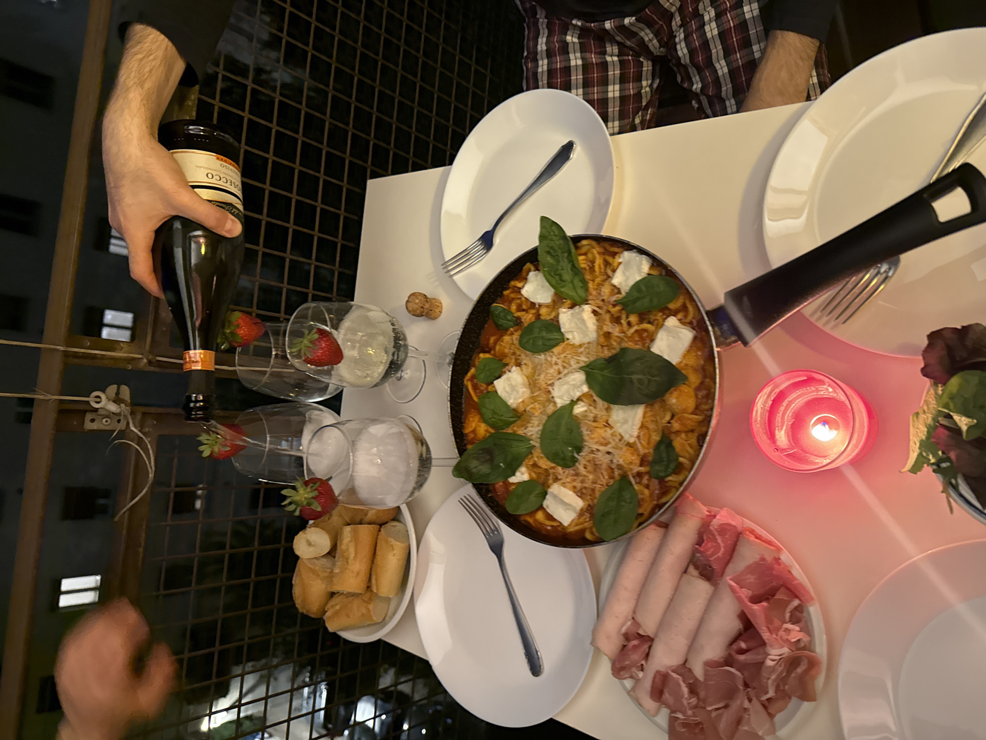 An overhead photograph captures a cozy evening dinner scene set on an outdoor balcony in Milan, Italy. The table is laden with a variety of dishes and drinks, illuminated by the warm glow of a candle and distant city lights, signifying nighttime.
In the upper left portion of the image, a person's arm is seen holding a bottle of Prosecco, its label clearly displaying "PROSECCO," as if preparing to pour. Two wine glasses are arranged nearby, each containing ice and a fresh strawberry, with a third empty glass also holding a strawberry. Two small, round objects, possibly corks, rest near one of the wine glasses.
Central to the table is a large, dark pan filled with a pasta dish, likely rigatoni, tossed in a rich tomato-based sauce. It's generously garnished with dollops of white cheese (resembling ricotta or fresh mozzarella), vibrant green basil leaves, and grated cheese. To the right of the pasta, a white plate presents an assortment of cold cuts, including neatly rolled slices of ham or turkey and delicate, folded slices of prosciutto. A small bowl containing a green leafy salad is also visible. On the left side of the table, a bowl holds various slices of baguette or small bread rolls.
The table is set with several white plates and forks, indicating multiple diners. A prominent red glass candle holder, with a lit candle flickering inside, casts a soft, inviting light across the food. In the background, a metal railing suggests an outdoor or balcony setting, and blurred lights of urban buildings provide a nocturnal backdrop. The overall atmosphere appears relaxed and convivial, ideal for a pleasant evening meal.