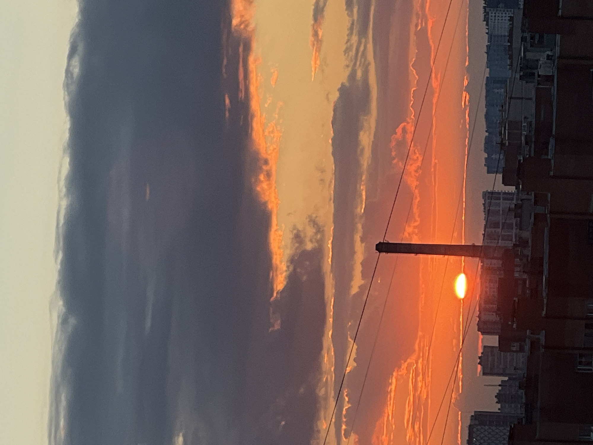 This is a vertical, eye-level media file of a sunset over a city. The sun is a bright orange orb low in the sky, partially obscured by clouds and a dark utility pole. The clouds are illuminated with vibrant shades of orange and yellow, contrasting with darker, grey clouds on the left side of the frame. The silhouettes of buildings line the right edge of the image, suggesting an urban environment. Power lines crisscross the scene, adding to the urban feel. The overall mood is serene and visually striking, capturing the dramatic beauty of a sunset. The time of day is clearly sunset, and the weather appears partly cloudy. There is no visible text in the media file.