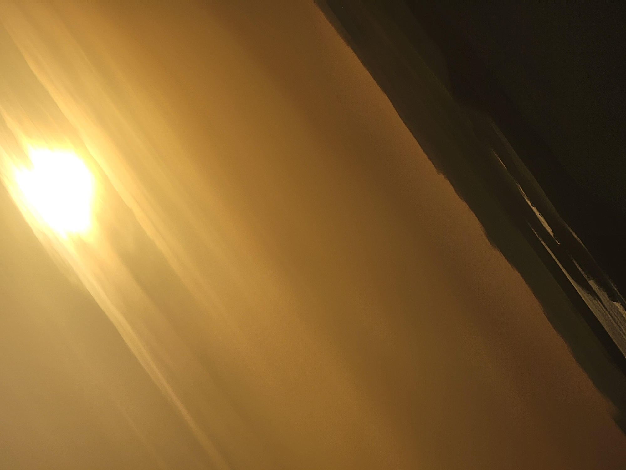 The image shows a hazy, golden sky with the sun prominently displayed in the upper left corner. Streaks of light radiate downwards and to the right. A dark, silhouetted landscape runs diagonally across the bottom portion of the image, appearing to be a sloping terrain or perhaps buildings. The overall impression is of a sunset or sunrise with a significant amount of atmospheric haze, possibly dust or smoke. The location is identified as Quarteira, Portugal.
