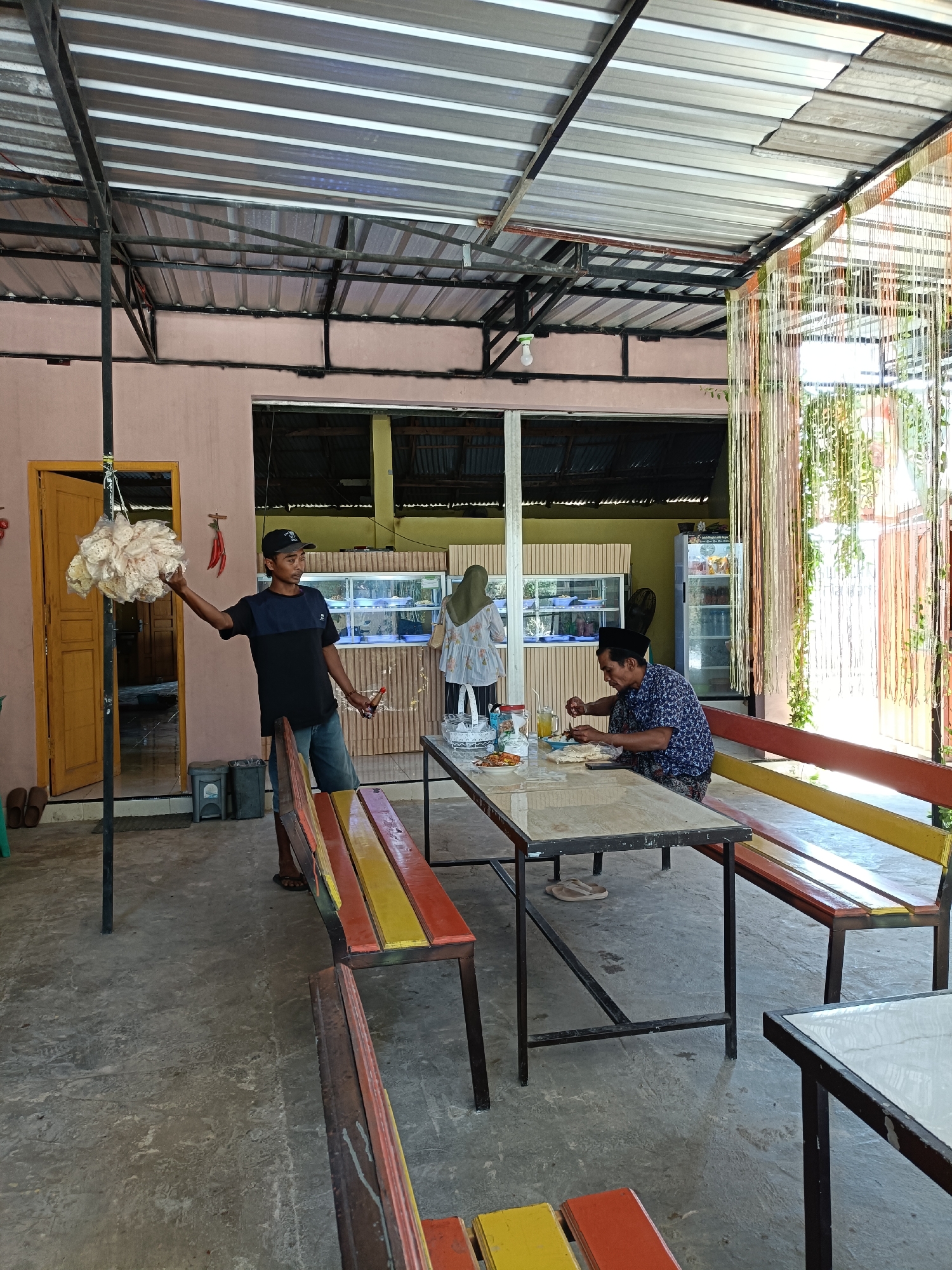 The image depicts an outdoor eating area at a casual restaurant in Pordapor, Indonesia. A young man in a black and blue t-shirt and denim shorts stands near a yellow door, holding a large bunch of white fried crackers. He appears to be a vendor or staff member. Two men are seated at a long, rectangular table. One man, wearing a blue and white patterned shirt and a black cap, is eating. The other man, wearing a blue and white patterned shirt and a black cap, is also eating. A woman wearing a light-colored headscarf and a patterned top stands in the background, looking towards a display case. The environment includes concrete flooring, a corrugated metal roof supported by dark metal beams, and colorful wooden benches with red, yellow, and orange slats. Decorative string lights hang from the roof. In the background, a refrigerator and display cases are visible. The overall scene suggests a relaxed, communal dining atmosphere.