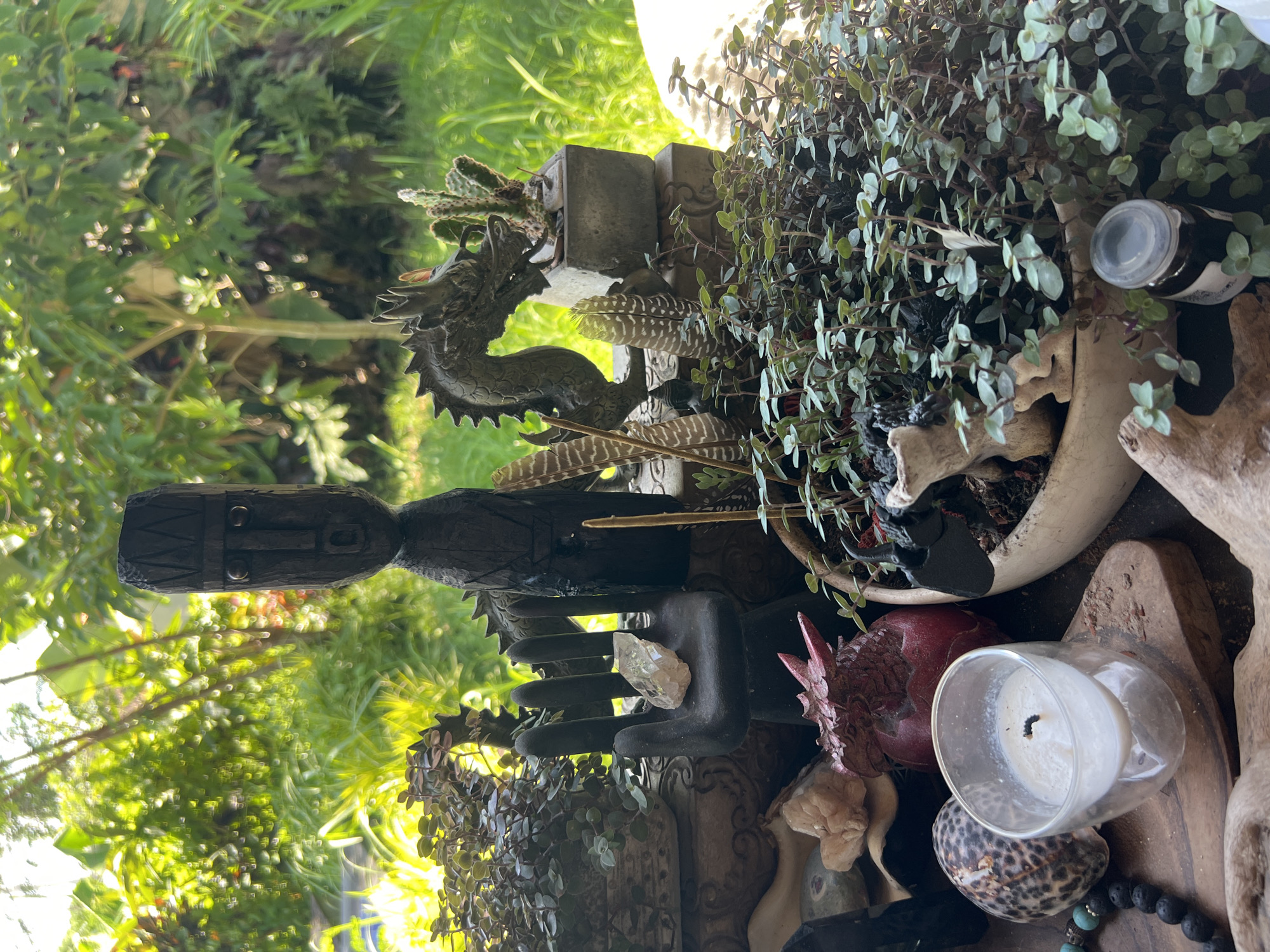 The image depicts an outdoor still life. Prominent elements include a tall, dark wooden figure with carved features, a bronze-colored dragon statue, and a hand-shaped sculpture holding a quartz crystal. Several feathers and small potted plants with dark green leaves are arranged around these central items. A lit candle in a clear glass holder rests on a wooden surface, alongside a leopard-print shell, a red pomegranate-shaped object, and what appears to be a dark stone beaded bracelet. The backdrop features lush green foliage. Based on the presence of specific decorative items (wooden figure, dragon), this image may be taken in Sukawati, Indonesia, which is known for its art market.