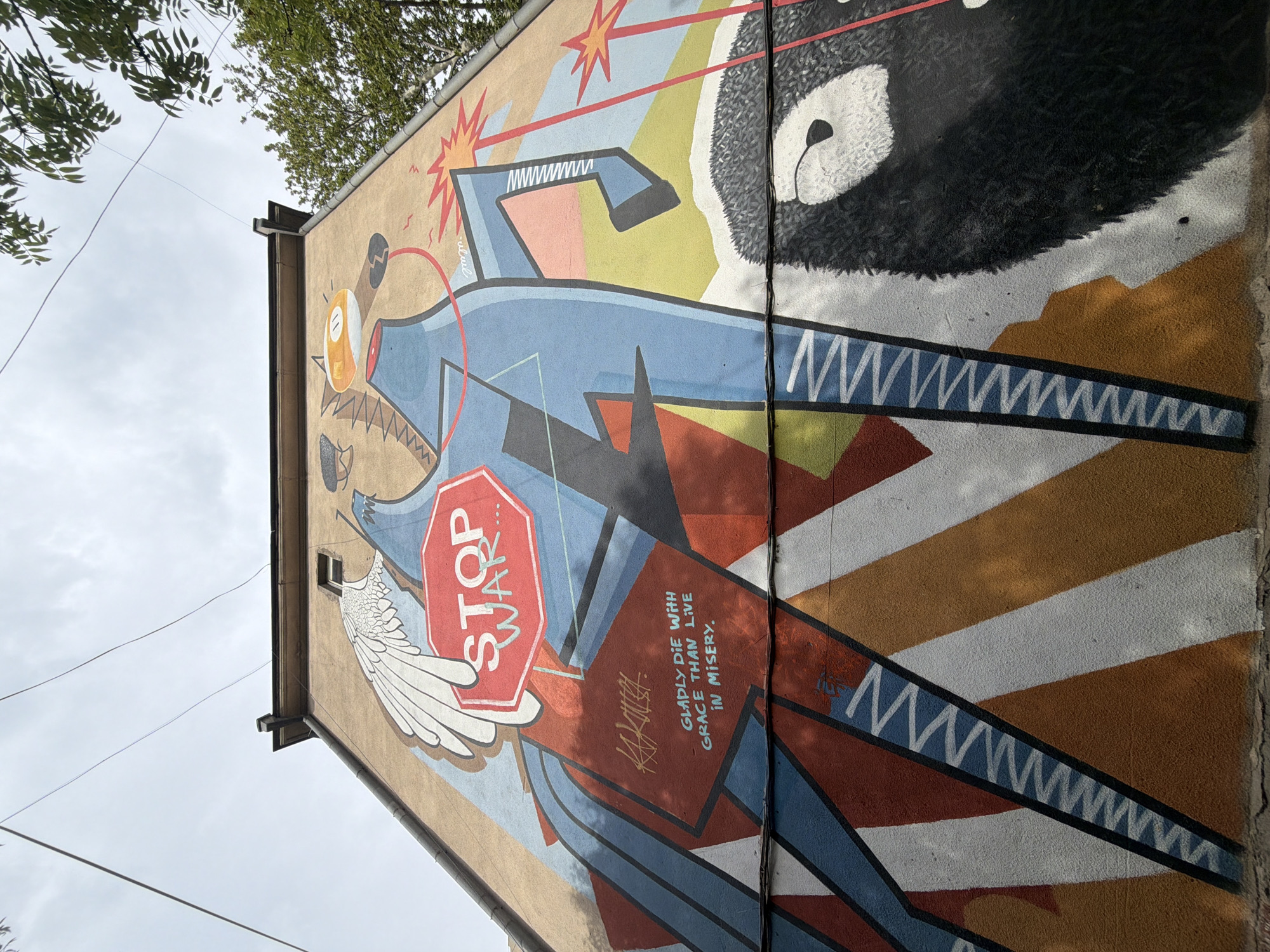 This media file captures a large, vivid mural painted on the side of a building in Tbilisi, Georgia, viewed from a low angle against an overcast sky and tree branches.
The mural depicts several stylized, dynamic figures. A central figure, appearing somewhat robotic or armored, holds a prominent red octagonal sign that boldly declares "STOP WAR" in white capital letters. Beneath this sign, a creature with outstretched white wings, resembling a bird or pigeon, is visible. On the upper right, a large, dark-furred creature, possibly a bear, with a distinctive white snout and eye, dominates the composition. Various abstract shapes and patterns in blues, yellows, reds, and browns form the background, punctuated by stylized starbursts or explosions.
The powerful anti-war message is reinforced by text painted below the "STOP WAR" sign, reading: "GLADLY DIE WITH GRACE THAN LIVE IN MISERY." This quote is written in a flowing, artistic script, accompanied by a signature or tag that resembles "KAZIKIAH."
The scene conveys a strong sense of protest and resistance. The building's architectural elements, such as a window, drainpipe, and the edge of the roof, frame the artwork. Utility wires are visible across the cloudy sky, placing the mural in an urban environment. Despite the overcast conditions, shadows suggest it is daytime. The vibrant colors of the mural contrast with the muted tan of the building wall.