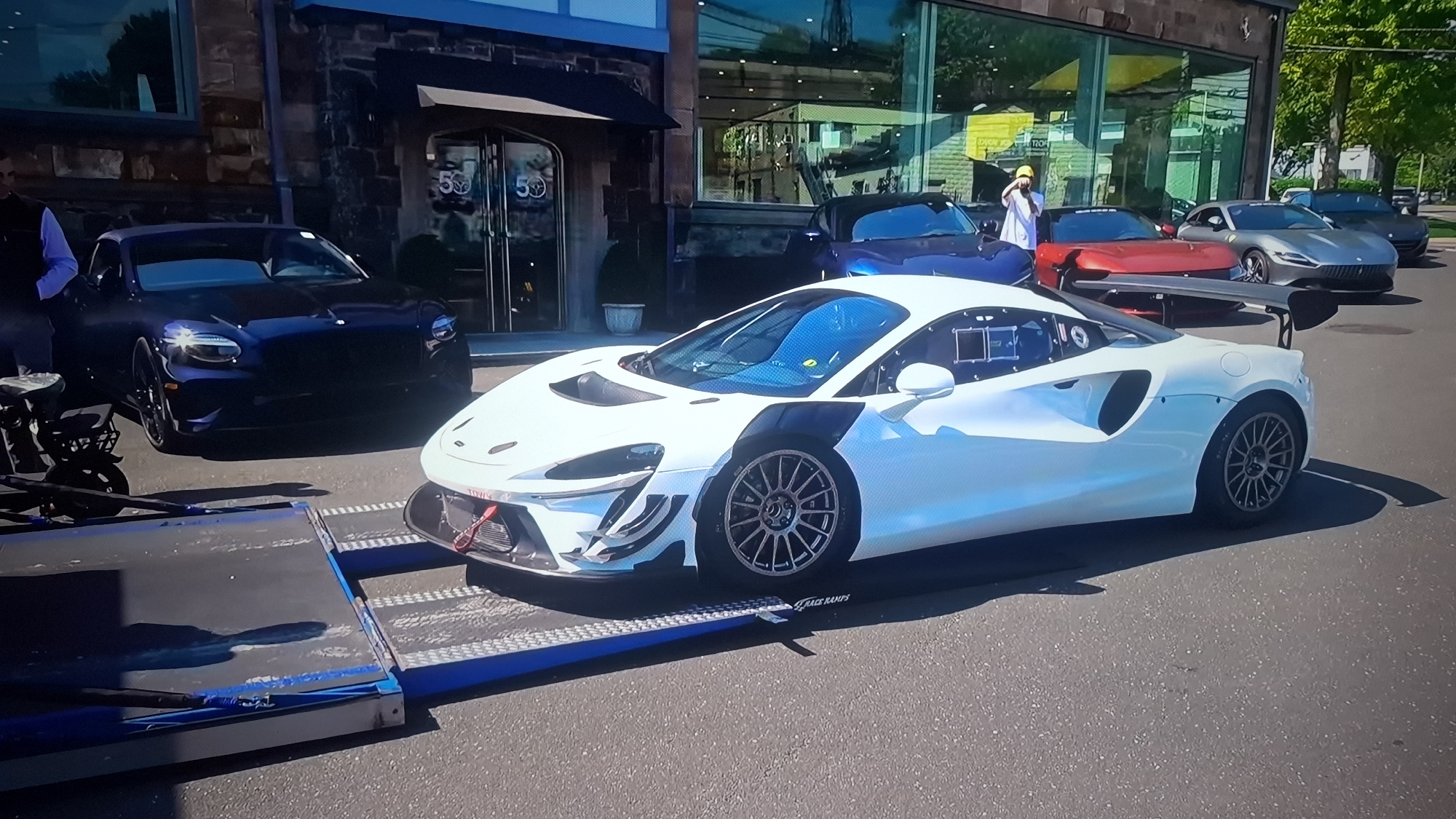 Here is a detailed description of the image:
**Scene:**
The image captures a street scene in what appears to be an affluent area of a city. The location is Unknown, Unknown.
**Subjects and Objects:**
* **Vehicles:** Prominently featured are several high-end cars. A white McLaren race car is on a car trailer and is the primary subject. Other cars include a dark blue convertible, a red sports car with a large wing, and two gray cars, one of which resembles a Ferrari Roma.
* **People:** At least two people are visible. One person is standing near the red car, wearing a white coat and a yellow hat and holding what appears to be a phone to their ear. Another person is near the blue convertible.
* **Buildings:** A stone building with a glass front is in the background. The building's doors have "50" displayed on them. The facade of the building is of stone construction with some dark accents.
* **Other:** A trailer and a vehicle transporter are positioned in the foreground, supporting the McLaren race car.
**Actions and Interactions:**
* The McLaren is being transported on a trailer.
* The person near the red sports car appears to be on the phone and is possibly taking a photo.
* The presence of multiple luxury vehicles suggests a car meet or a showroom event.
**Environment:**
* **Setting:** The scene is set on a paved road with the vehicles parked or being transported.
* **Weather/Lighting:** It is daytime with ample sunlight casting shadows on the road and the cars.
* **Context:** The scene suggests a showcase or event related to luxury and performance vehicles.