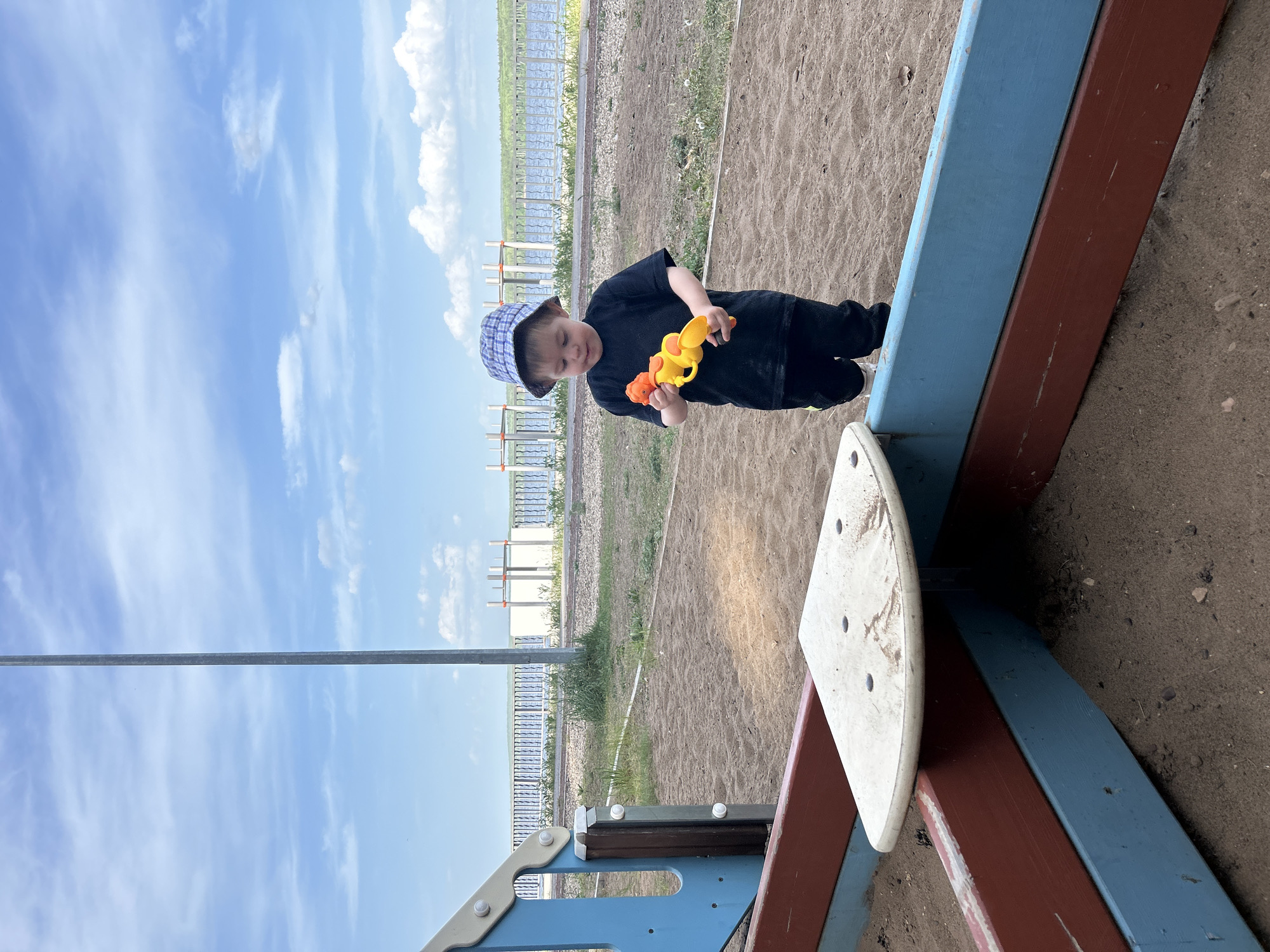 A toddler in a dark shirt, dark pants, and a plaid hat stands on a playground, holding a yellow and orange toy. The playground equipment is constructed of blue and brown wood. The ground is covered in sand. Behind the playground, there is a fence, grass, and a body of water. The sky is blue with scattered clouds. The image was taken outside on a sunny day. The location is Chaikovsky, Russia.