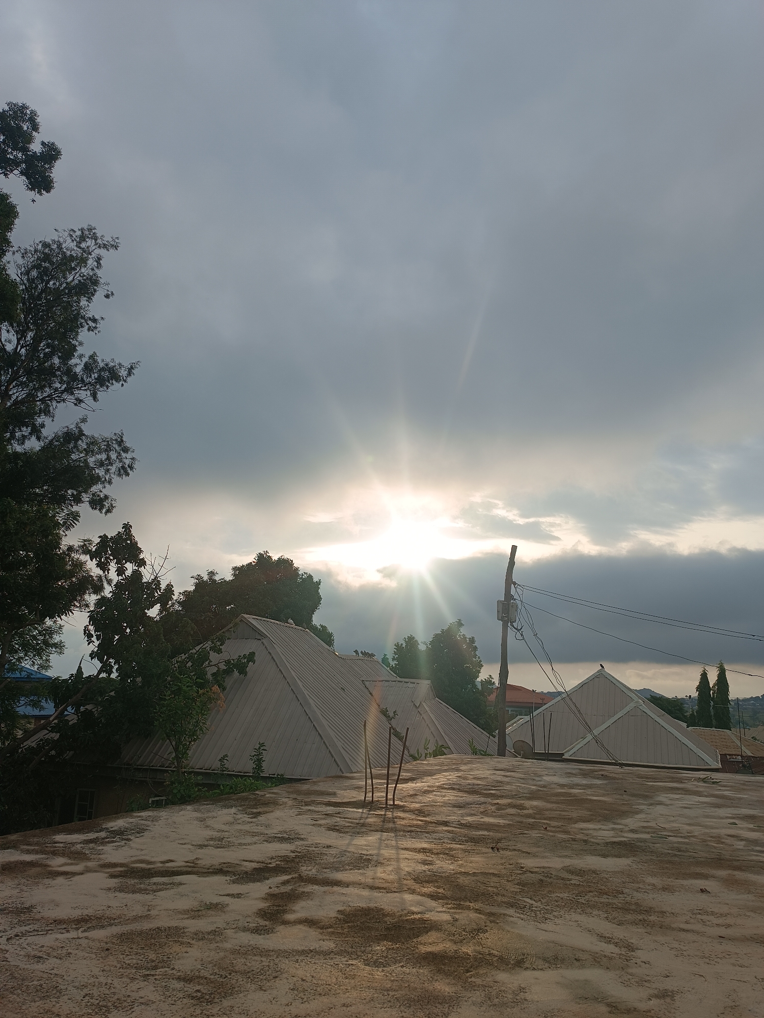 The image captures a serene, partly cloudy scene from a rooftop perspective in what is contextually known as Bukuru, Nigeria. The sun, positioned prominently to the left of the center, breaks through a layer of dark gray clouds, casting brilliant rays and creating a strong lens flare effect. The sky transitions from bright near the sun to a darker, overcast appearance towards the edges of the frame.
In the foreground, a weathered, flat concrete rooftop dominates the lower portion of the image, displaying a textured surface with irregular dark stains and patches. Beyond this roof, several buildings with light-colored, steeply pitched, corrugated metal roofs are visible. Some of these roofs feature distinct gable ends. Lush green trees, with dense foliage and visible branches, frame the left side of the image and are interspersed among the buildings in the middle ground, adding to the natural elements of the scene.
A tall, weathered wooden utility pole, equipped with a small gray box and multiple dark electrical wires, stands in the middle-right section of the image. The wires extend horizontally across the frame. A small, dark bird is perched atop the peak of a gabled roof to the right of the utility pole. In the far background, faint outlines of distant hills or mountains can be discerned under the cloudy sky.
The strong sun rays and the angle of the light suggest either early morning or late afternoon, possibly signaling a sunrise or sunset. The overall atmosphere is quiet and somewhat dramatic due to the interplay of light and shadow from the sun and clouds. No visible text is present in the image.