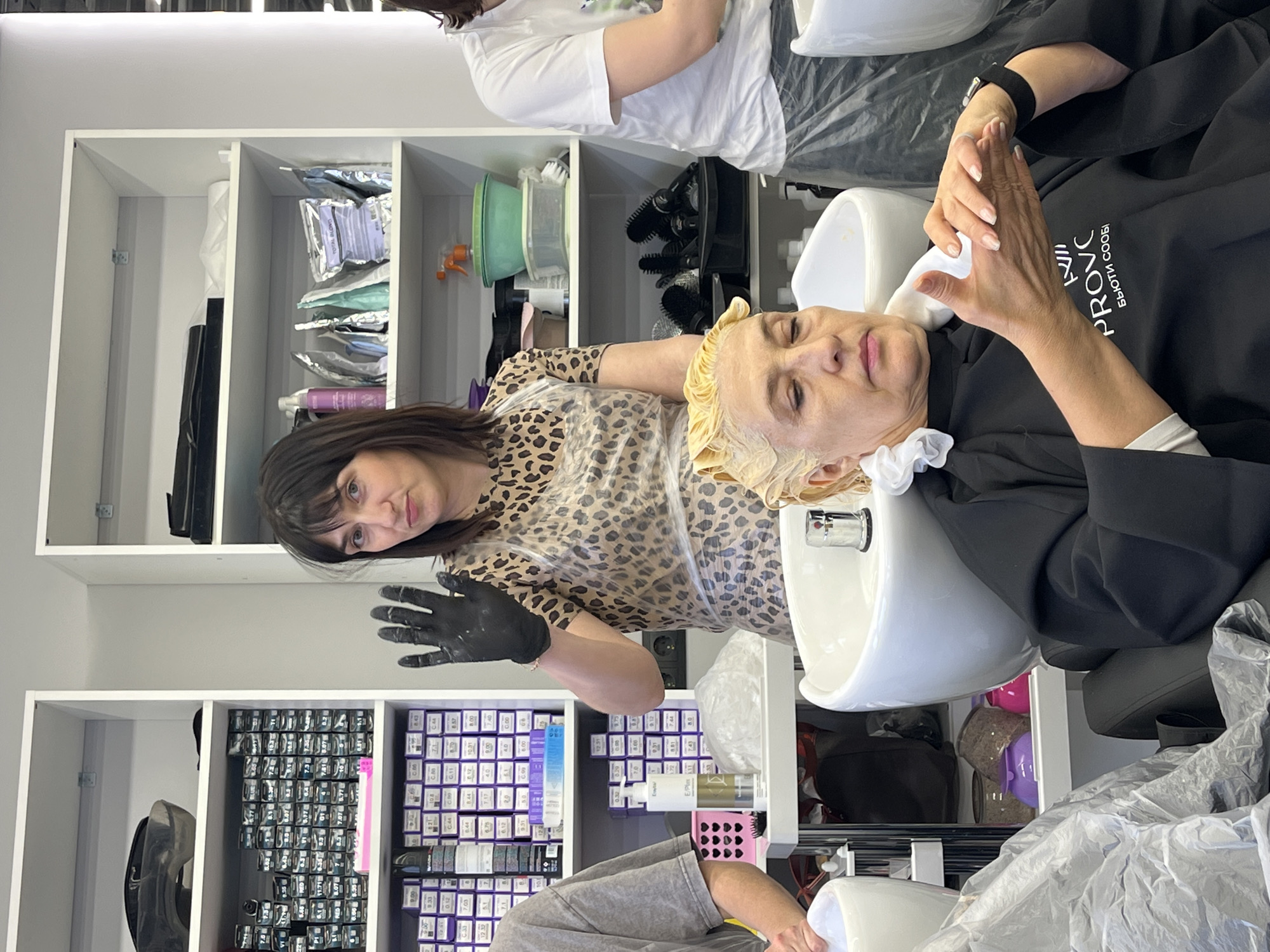 The image captures a hair treatment in progress within a well-lit hair salon located in Izhevsk, Russia.
**Subjects:** A female client, likely in her 50s or 60s, is seen reclining in a black salon chair with her head over a white hair washing basin. Her eyes are closed, suggesting a relaxed state. A female stylist, wearing a leopard-print top and black gloves, stands beside her, looking towards the camera. Part of another person's legs and lower torso are visible in the bottom foreground, seated in another salon chair.
**Activity or event taking place:** The client is undergoing a hair treatment, most likely coloring or bleaching, as indicated by the yellow processing cap on her head and the light-colored product visible on her hair. The stylist's black gloves suggest she is actively engaged in applying or rinsing chemical treatments.
**Objects:** Prominent objects include a white ceramic hair wash basin, salon chairs, a black protective cape worn by the client, a yellow hair processing cap, and a wide array of hair care products neatly arranged on white shelves in the background. These products include numerous boxes of hair dye (with numerical codes), bottles, a green mixing bowl, and a spray bottle.
**Notable details:** The salon appears clean and organized, with a clear focus on hair treatment services. The client's posture and closed eyes convey a sense of calm or relaxation. The stylist's professional attire and gloves indicate a skilled service. The time of day is indeterminate due to the indoor setting with artificial lighting.
**Visible text:** On the client's black protective cape, white text reads "PROVC" and "БЬЮТИ СООБ," which translates to "BEAUTY COMMUNITY." Various numerical codes, indicative of hair dye shades (e.g., 7.0, 9.1, 10.0, 9.32), are visible on product boxes on the shelves. A faint "Estel E/Plus" can be read on one of the product bottles.