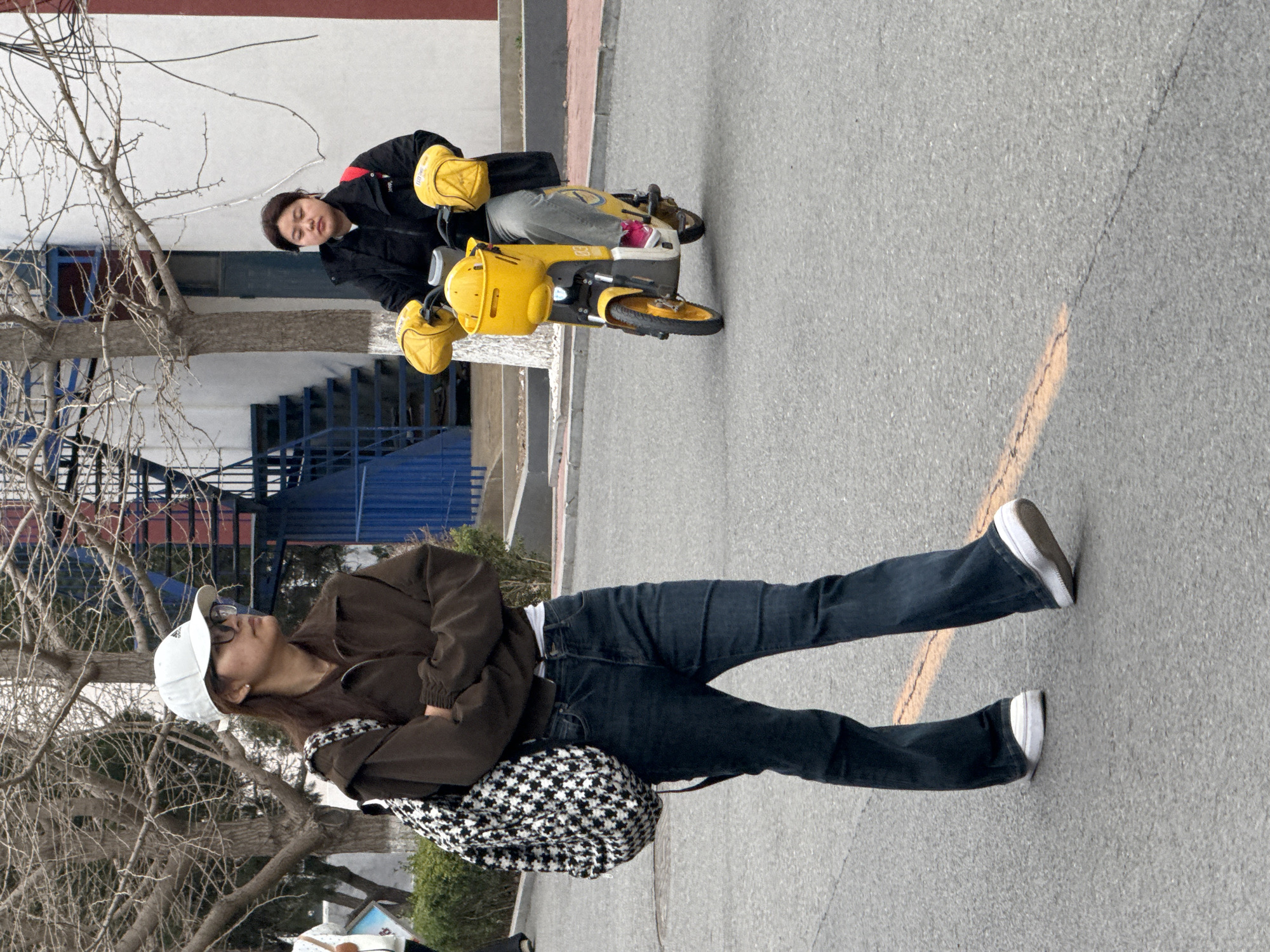 Two people are on a paved road. One person is standing wearing jeans, a brown jacket, a white hat, and a checkered backpack. The other person is sitting on a yellow scooter and wearing a black jacket and yellow helmet. In the background, there is a building with blue stairs and bare trees. The scene is outdoors on a sunny day. The location is in the city of Zhongtun, China.