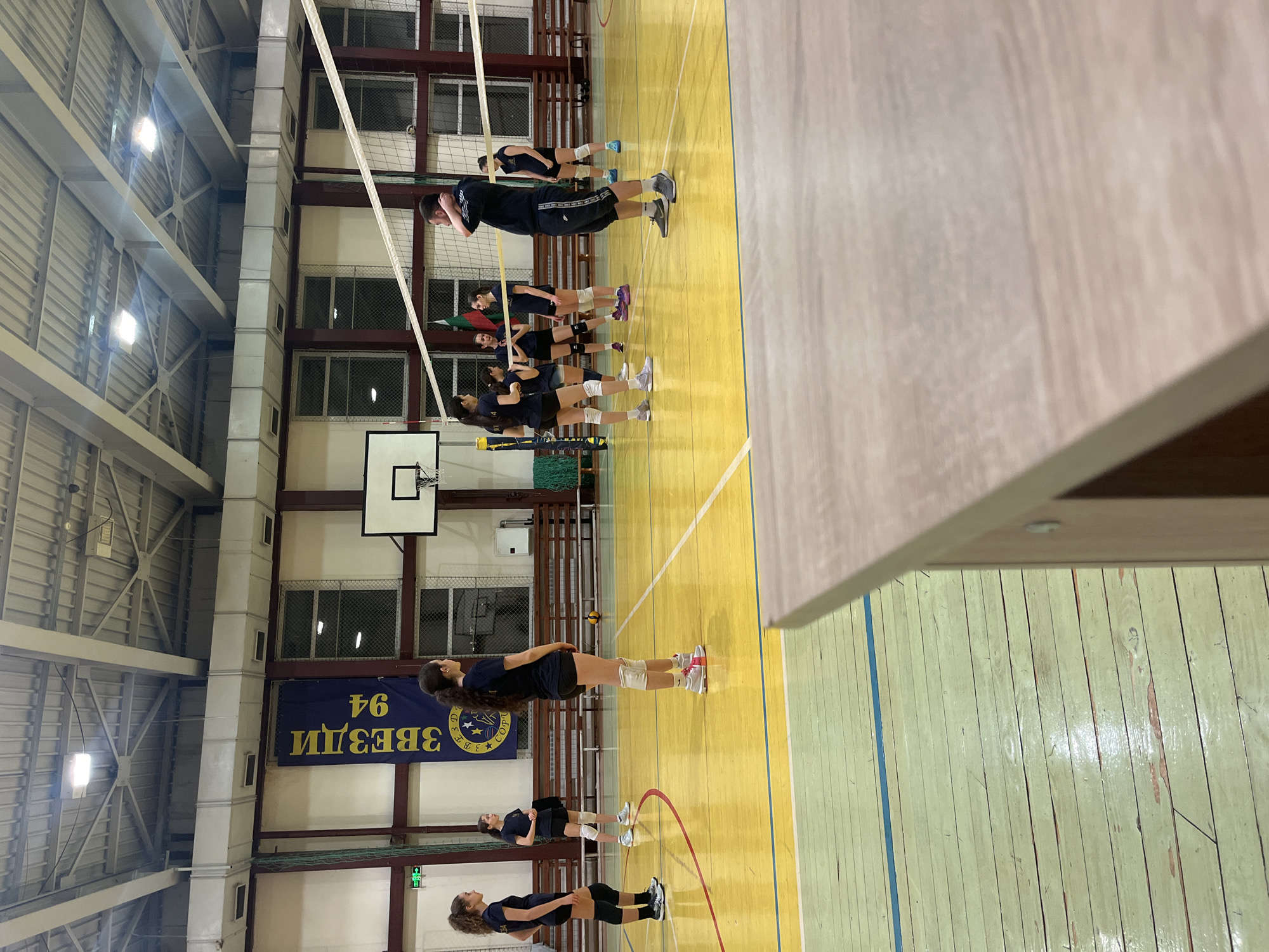 A volleyball team is lined up on the court of a gymnasium. The players are wearing dark shorts and shirts, and some have knee pads. A net is strung across the court, and a banner hangs on the wall behind them. The banner reads "ЗВЕЗДИ 94," which translates to "Stars 94" in English. The scene is likely set in Sofia, Bulgaria, given the Cyrillic lettering on the banner.