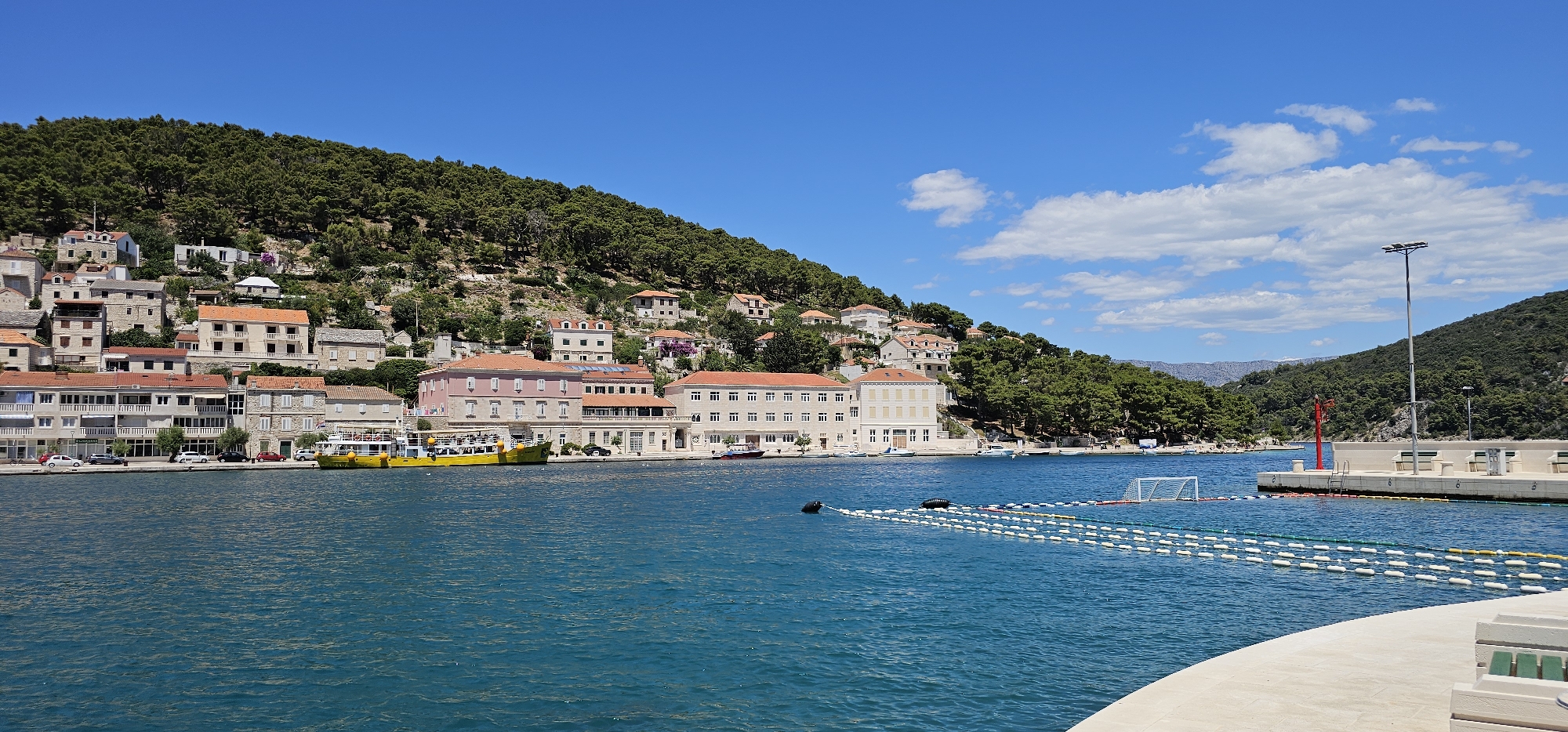 This is a wide-angle, eye-level photograph of a harbor in Općina Pučišća, Croatia. The image is dominated by the vibrant blue water of the sea in the foreground, which occupies the lower half of the frame. A concrete promenade curves into the lower right corner of the image, with white bench seating visible. In the mid-ground, a yellow ferry is docked, its deck lined with passengers. Behind the ferry, a row of multi-story buildings with terracotta roofs line the waterfront. The buildings are a mix of architectural styles, with some featuring stone facades and others painted in pale colors. Behind the buildings, a densely forested hillside rises steeply, covered in dark green pine trees. The sky is a clear, bright blue with a few wispy white clouds scattered across the upper portion of the frame. A tall lamppost stands on the right side of the harbor, near a red marker buoy and a floating water polo goal. The overall impression is one of a sunny, idyllic coastal town on a clear summer day.