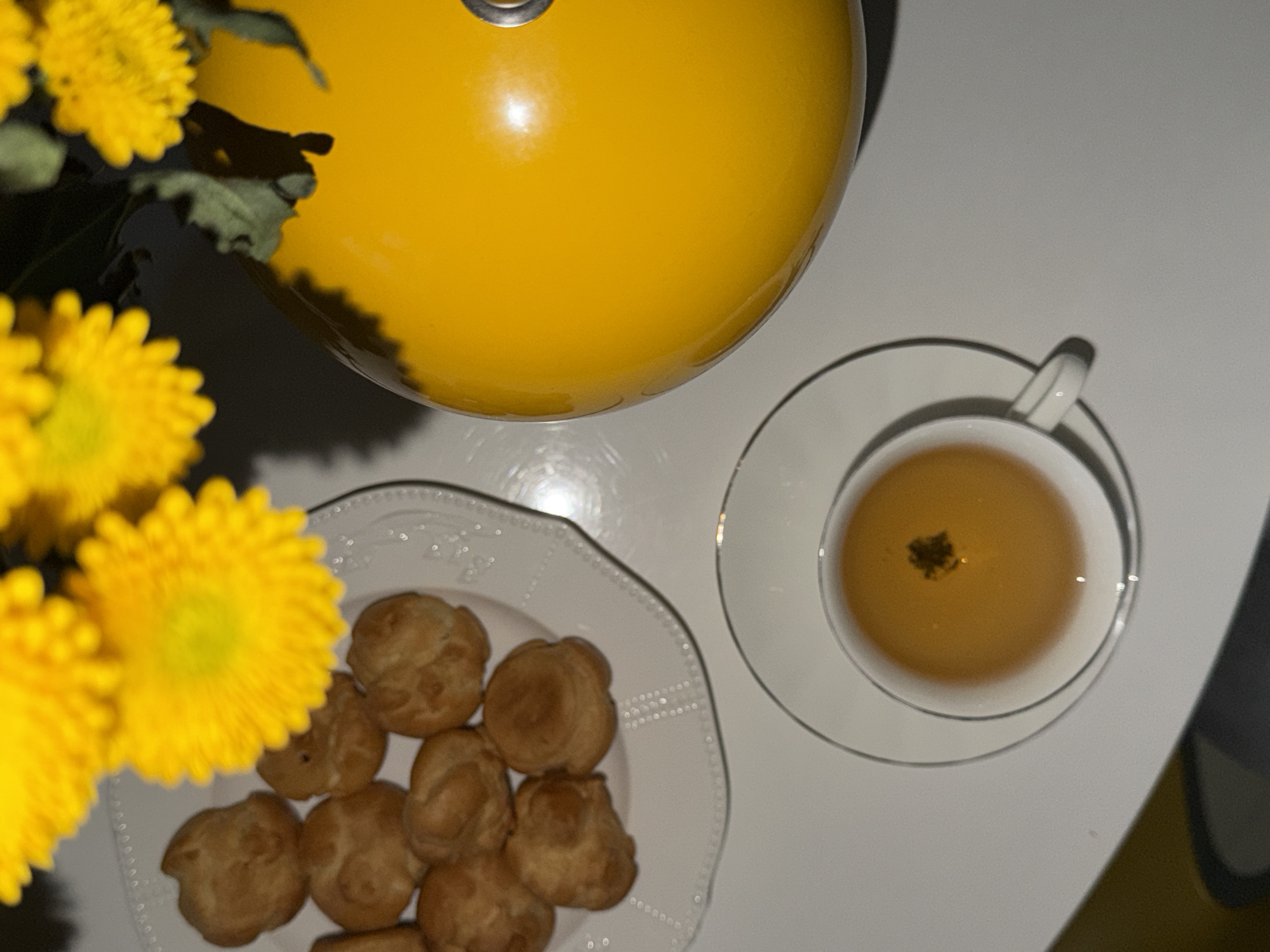 A top-down shot shows a white surface with a yellow teapot, a white teacup and saucer filled with amber liquid, a white plate with small pastries, and yellow chrysanthemums in the lower left corner. There is no evidence to support the presence of Khimki, Russia. No people, subjects, actions or interactions are visible.