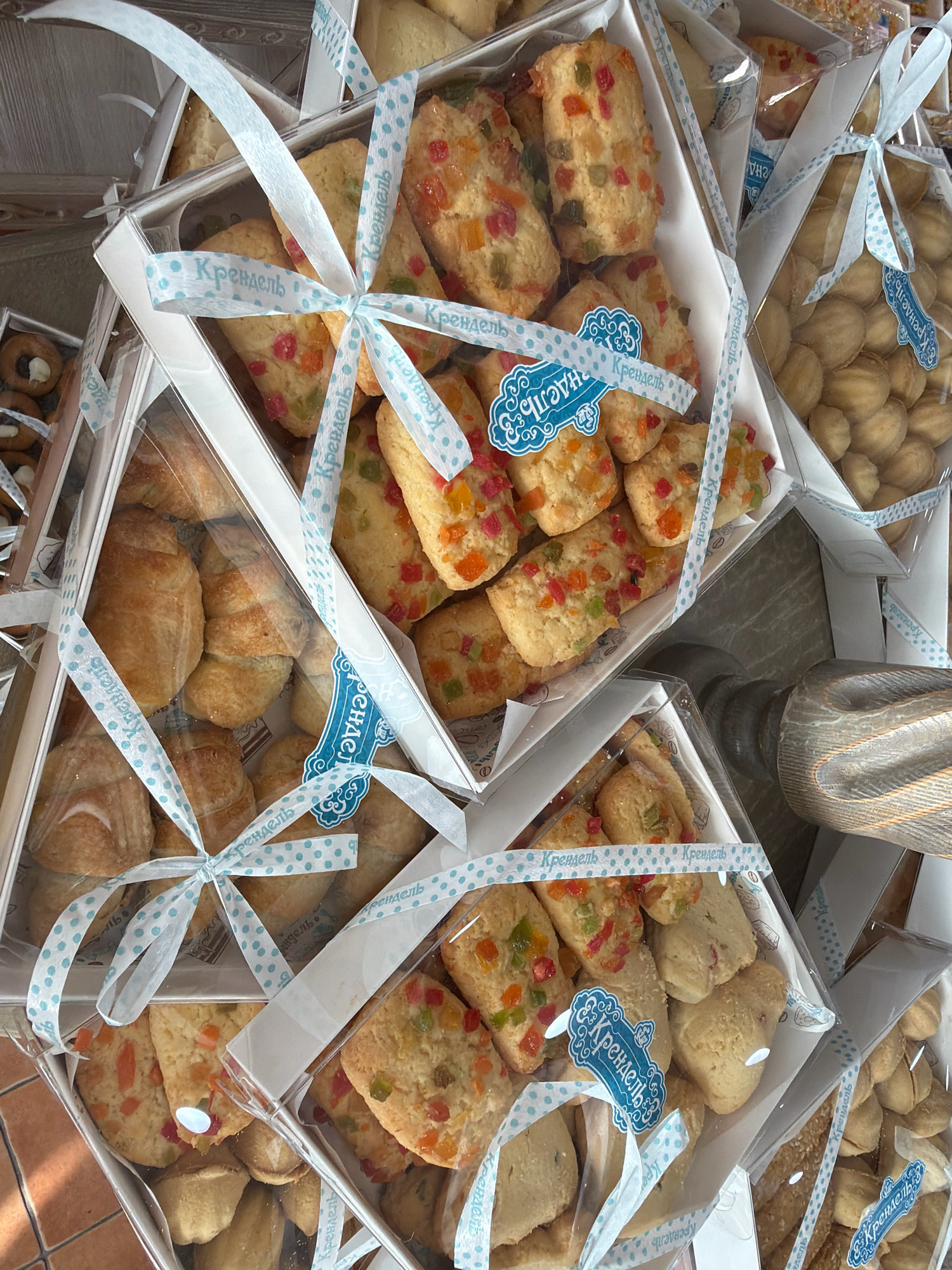 The image displays a close-up view of several transparent boxes filled with assorted baked goods, likely cookies and pastries, tied with light blue polka-dot ribbons. The boxes are stacked and arranged in a manner suggesting they are for sale or display. Some of the cookies appear to be shortbread or similar, studded with colorful candied fruit pieces. Other boxes contain what look like small croissants or rolls. In the background, a wooden object with a textured surface is visible, possibly a table or a decorative element. The overall setting seems to be an indoor retail environment, such as a bakery or a confectionery shop. No people are visible in the frame. The text on the ribbons is in Cyrillic script and reads "Крендель" (Krendel), which translates to pretzel or roll, suggesting the brand or type of product. Based on the given information, the image is from Pavlodar, Kazakhstan.
