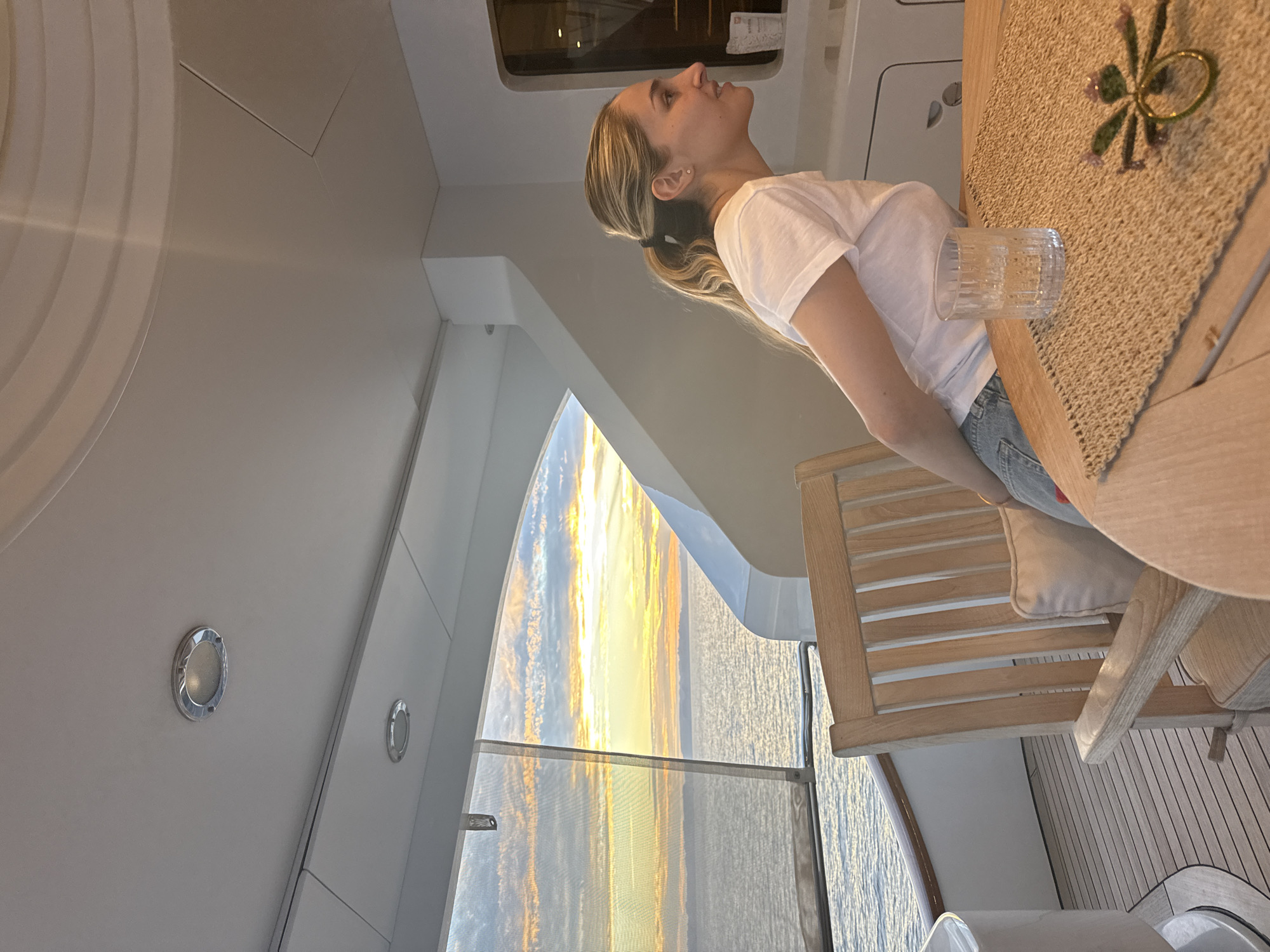 A woman sits at a table on a yacht, overlooking the ocean, presumably near Maravia, Mexico. The woman has blonde hair tied in a ponytail, wearing a white t-shirt and jeans. She is looking up and away from the camera. On the table is a woven placemat, a clear glass of liquid, and a glass floral decoration. A wooden chair supports the woman. Outside a large window is a sunset over the ocean. The yacht interior is modern with white paneling and wood flooring.